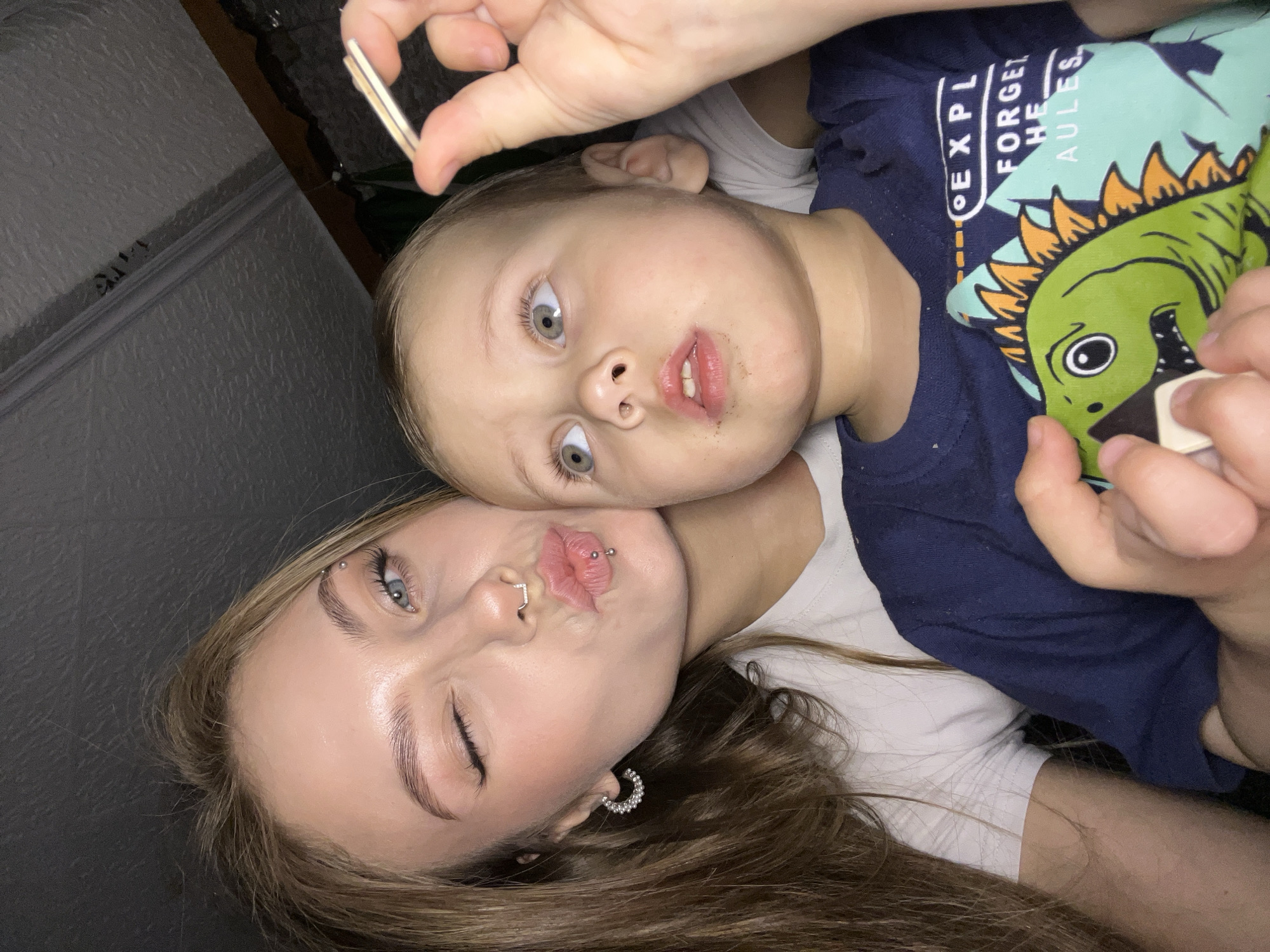 A woman and a young child are lying down close together, taking a selfie. The woman has light brown hair, fair skin, a nose ring, and an earring. She is winking at the camera with her lips puckered. The child has fair skin and light hair. He is wearing a blue shirt with a green dinosaur on it. The background appears to be a wall with textured wallpaper. They are holding a small brown and white object. The location in the city of Фоки, Russia is not determinable from the image.