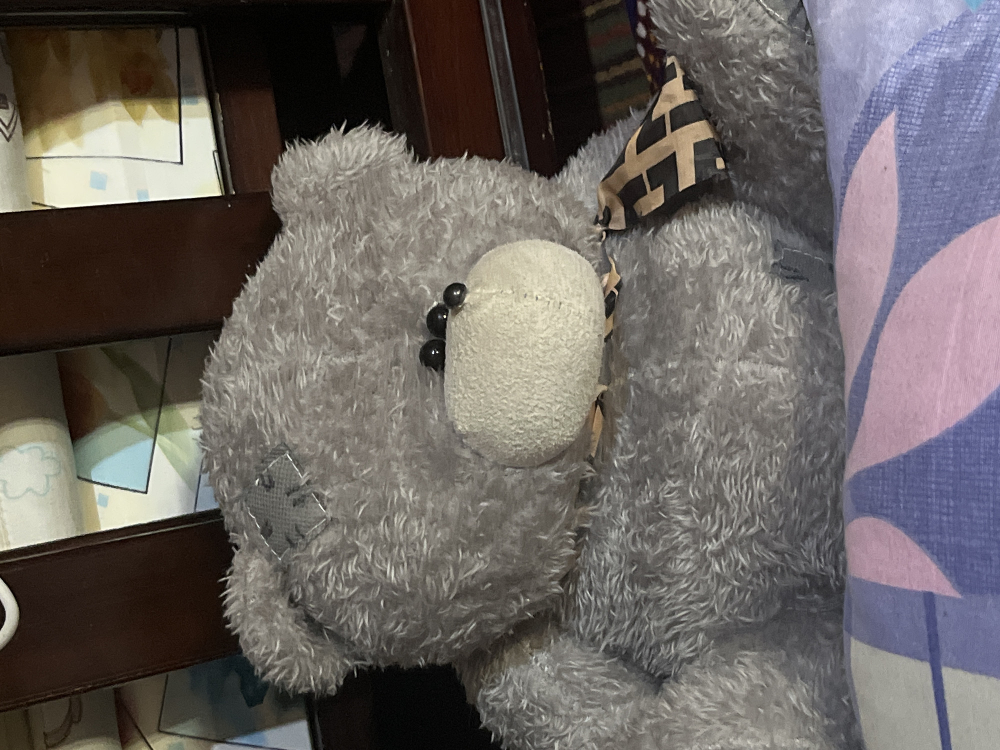 A grey teddy bear with black eyes and a patterned bandana is seated on a surface with a pink and purple patterned fabric in what appears to be a bedroom or nursery setting. The bear is plush and has a somewhat worn appearance, with a small grey patch on its forehead. The background consists of wooden furniture and what looks like a crib or bed with colorful bedding. There is no visible indication of the city of Kuyun, Uzbekistan.