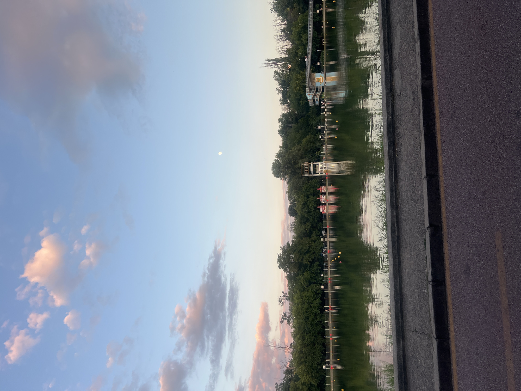 This image depicts a serene waterscape in Plovdiv, Bulgaria, likely at the Plovdiv Rowing Canal. The foreground shows a paved walkway with a concrete curb, bordering a stretch of asphalt road marked with a yellow line. Beyond the walkway lies a body of water, reflecting the sky and surrounding greenery. In the middle ground, a row of trees forms a lush green backdrop along the far bank of the canal. Scattered on the water are yellow buoys. A diving platform with white railings and a white structure is visible on the left, and a colorful building with blue and orange accents, featuring stairs and a walkway, is present on the right. The sky above is a gradient of pale blue, transitioning to lighter hues near the horizon. Wispy clouds, tinged with pink and orange, are scattered across the upper portion of the sky, suggesting either sunrise or sunset. A faint full moon is visible in the clear sky on the right side of the image. The overall atmosphere is tranquil and picturesque, capturing a moment of calm by the water.