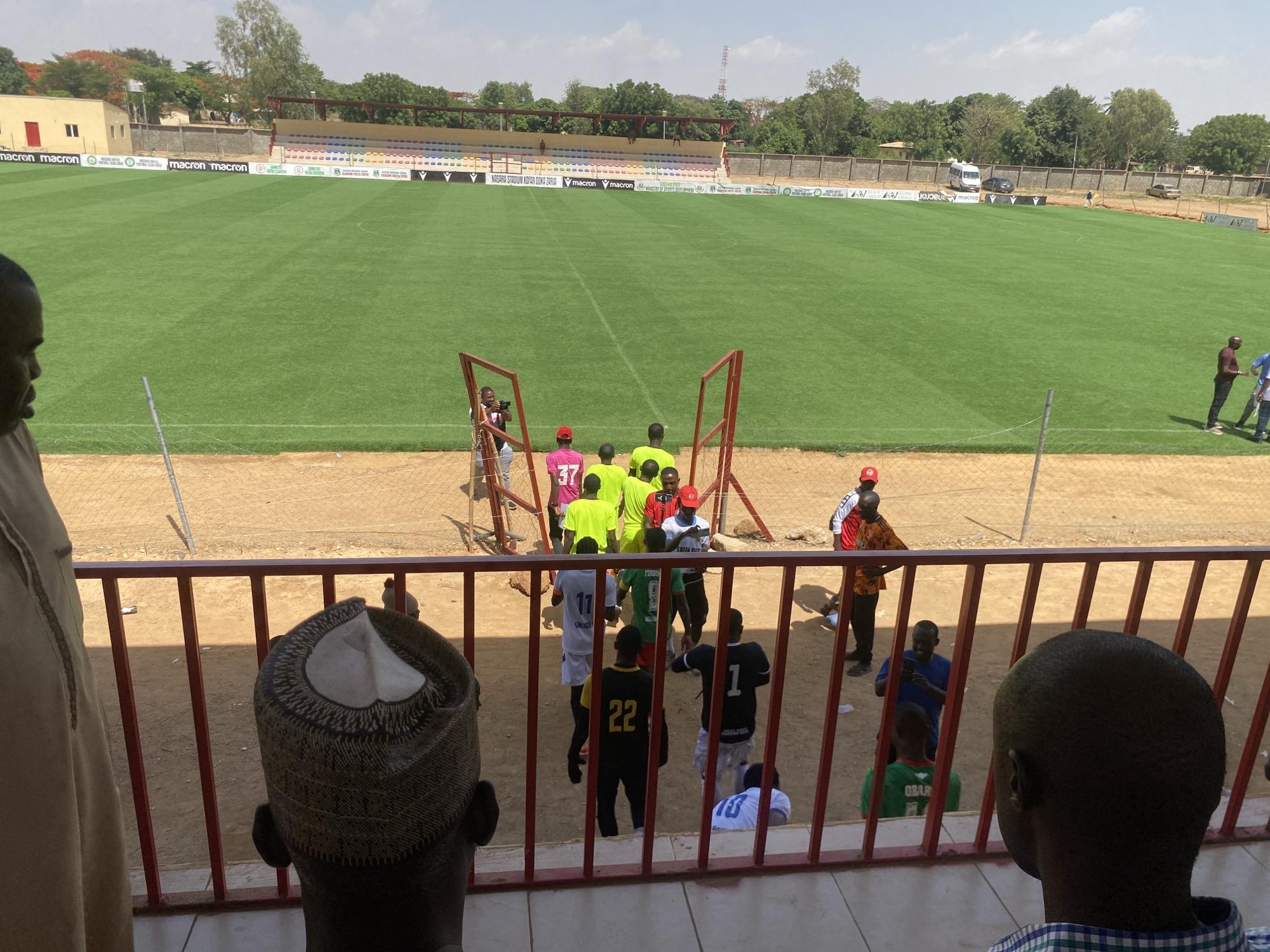 The image shows a soccer field at Nasarawa Stadium in Tudun Wada, Nigeria. A group of people are gathered near one of the goals, wearing various jerseys and casual clothing. A few people are standing behind a red railing, looking at the field. In the background, a stadium with rows of seats is visible, with "Macron" branding displayed. The weather appears sunny and clear. Buildings and trees line the perimeter of the stadium.