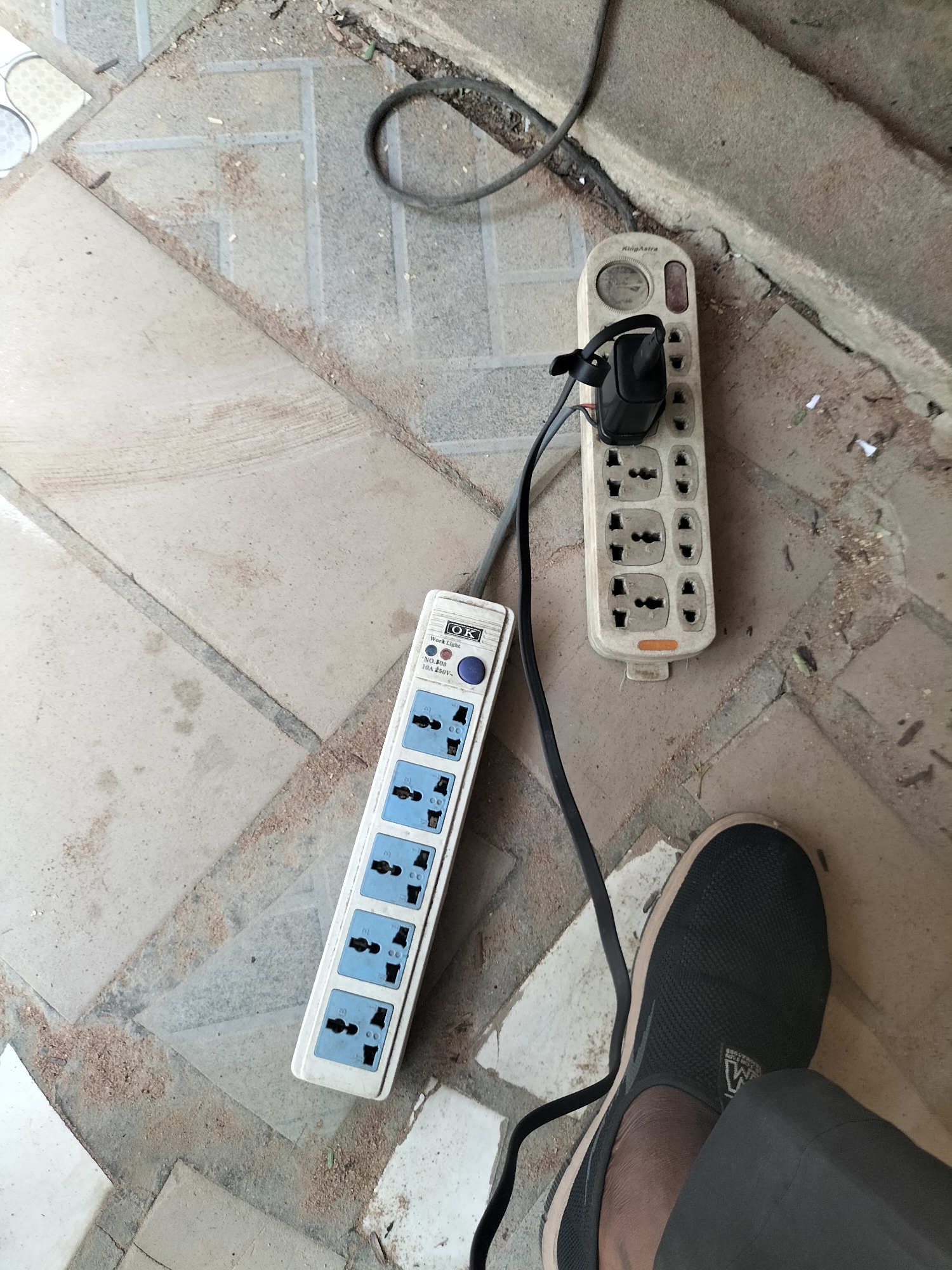 The image shows two power strips on a tiled floor. The floor is composed of light-colored square tiles with darker grout lines, some of which are cracked. There are scattered pieces of debris, including what appears to be wood shavings and small white paper fragments, on the floor.
One power strip, labeled "KingAstra," is white and has multiple outlets. A black electrical plug is inserted into one of the outlets and secured with a black tie. A dark gray electrical cord extends from this power strip and is partially coiled on the floor.
The second power strip is also white, with blue outlets, and is labeled "OK" with "Work Light" and "NO.808 10A 250V~" printed on it. This power strip is positioned to the left of the KingAstra strip and has multiple outlets, all of which appear to be empty. A dark gray electrical cord connects to this power strip and runs along the floor, appearing to join the cord from the other power strip.
In the lower right corner of the image, the foot of a person wearing a dark gray sock-like shoe is visible, with a dark gray fabric garment next to it. The shoe has a white sole and a visible logo on the side. The person's lower leg is also partially visible.
The scene appears to be outdoors, possibly in a workshop or storage area, given the presence of debris and the type of electrical equipment. The lighting suggests it is daytime. The presence of the power strips and the electrical cord indicates a need for power. The location is identified as the city of Jalingo, Nigeria.