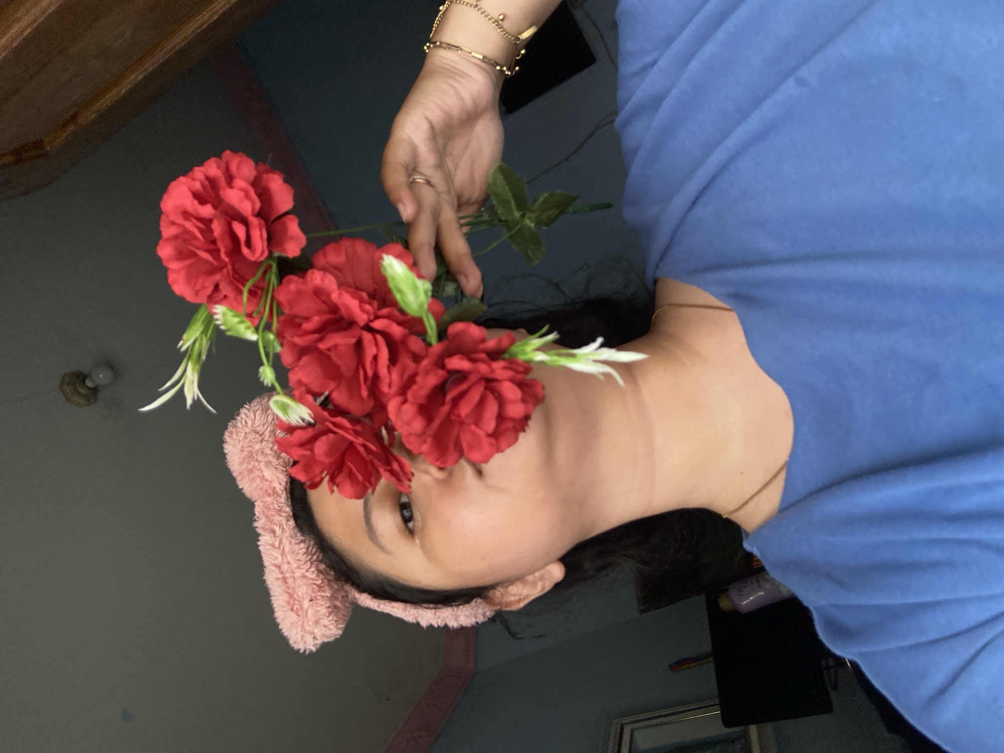 A close-up, portrait-style image features a person indoors, holding a bouquet of red artificial flowers to partially obscure their face. The individual is wearing a light blue t-shirt and a pink, fuzzy headband. Their right hand, adorned with multiple gold bracelets and a gold ring, holds the stems of the flowers, which have green leaves and small white flower buds. A thin gold chain is visible around their neck. The person's visible eye looks towards the camera with a neutral expression.
The background is dimly lit, predominantly featuring a dark wall. Visible elements include a wooden beam on the ceiling, a light fixture, and various household items such as a framed picture or mirror, dark furniture, and a small purple bottle. The setting appears to be a casual, residential indoor space. The location context is Gelam, Indonesia. There is no visible text in the image.