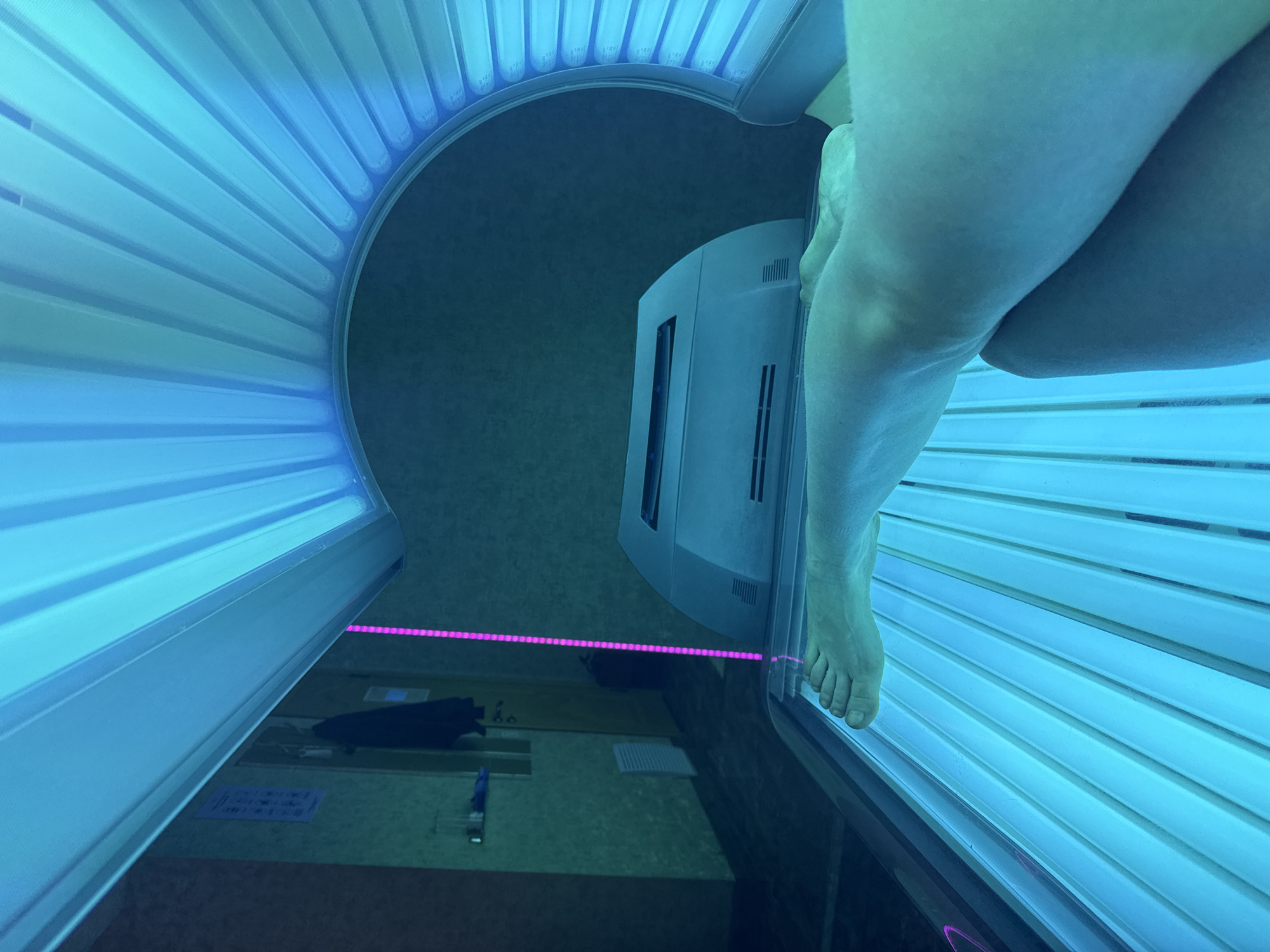 A person's legs and feet are visible in a tanning bed with blue lights. The tanning bed is open, and the lower half of a person is laying on it. There is a control panel near the head area and a pink LED strip on the left side of the tanning bed. The environment is indoors, possibly a tanning salon. The location is unknown and there's no visible information that suggests that the location is Saint Petersburg, Russia.