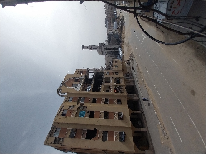 The image portrays a street scene in Khan Yunis, Palestinian Territory, marked by widespread urban destruction under an overcast sky. In the foreground on the right, dark electrical wires and a partially visible sign with Arabic text are present. A two-lane asphalt road, delineated by faded white markings, occupies the midground. Two individuals, clad in dark attire, walk on the sidewalk to the left of the road, moving away from the camera's perspective. Immediately adjacent to the sidewalk stands a multi-story building, tan with brown accents, exhibiting severe structural damage: large sections of its facade are missing, windows are shattered or absent, and a prominent arched architectural element on its upper floors is heavily compromised. A blue tarp hangs from a damaged window opening on a middle floor. Further down the road to the right, a mosque with a damaged minaret and a partially collapsed dome is visible, surrounded by extensive piles of concrete rubble and debris. Additional damaged structures and indistinct figures are present in the distant background.