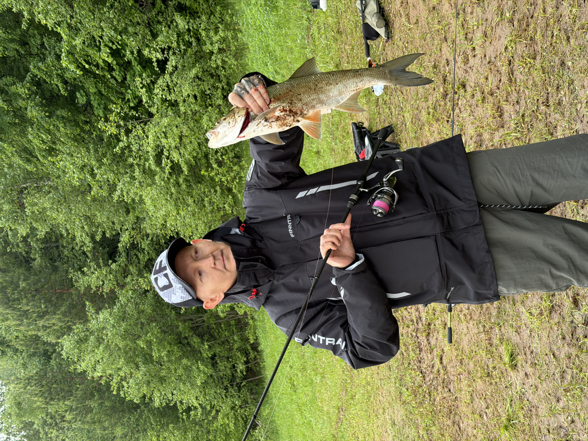 A man stands in a grassy area near trees, holding a large fish up with his left hand. He is wearing a black jacket with the word "FINNTRAIL" on the chest and "TRAIL" on the sleeve, grey pants, and a grey and white patterned baseball cap. He is holding a fishing rod with his right hand. There are trees and green vegetation in the background. The location is near Ольховка, Russia.