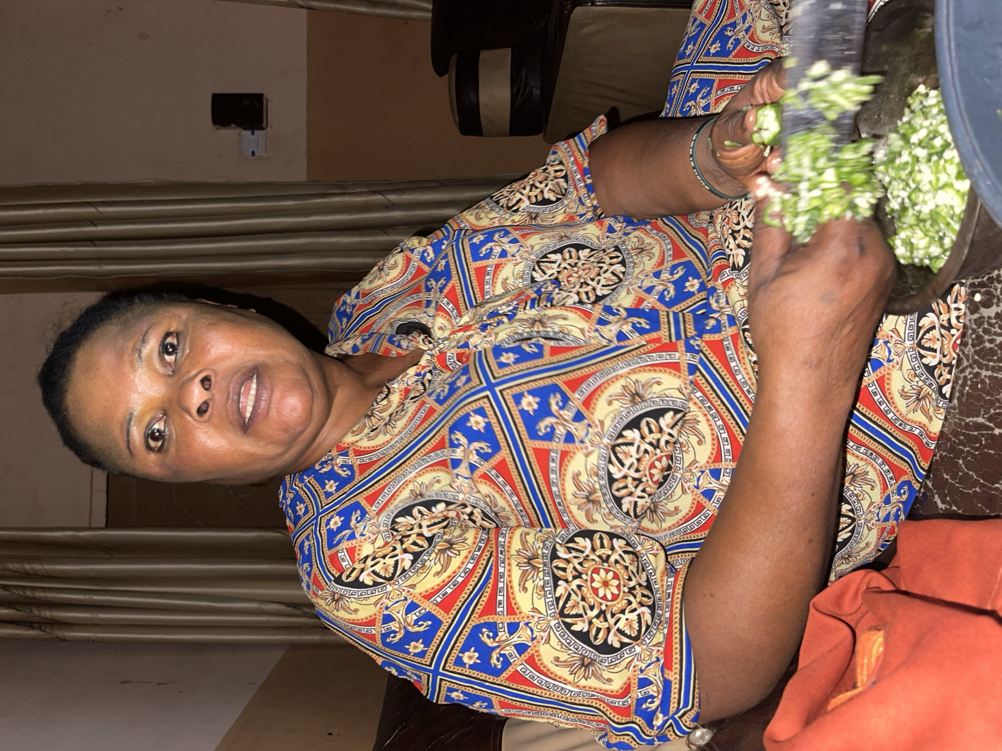 The image features an adult woman, likely of African descent, engaged in preparing food within what appears to be an indoor setting in Unguwar Gabas, Nigeria. She is positioned slightly to the left, looking directly at the camera with a gentle, amiable expression and a faint smile.
The woman has dark skin and short, dark hair. She is wearing a vividly patterned garment, possibly a dress or blouse, characterized by intricate designs in blue, red, yellow, and black. On her left wrist, she has multiple thin, green bangles. Her hands are actively involved in chopping or pounding green vegetables, which appear to be okra, using a dark utensil within a large, dark mortar positioned in front of her. The chopped vegetables are visible inside the mortar.
The background suggests a domestic interior. A light-colored wall with an electrical outlet or switch is visible behind her, along with light brown curtains or blinds. A dark brown piece of furniture, possibly an armrest of a sofa or chair, is partially seen on the left. In the lower right corner, a piece of orange fabric is also visible. The lighting in the scene is artificial and somewhat subdued, indicating an indoor environment, possibly during the evening. No text is visible in the image.