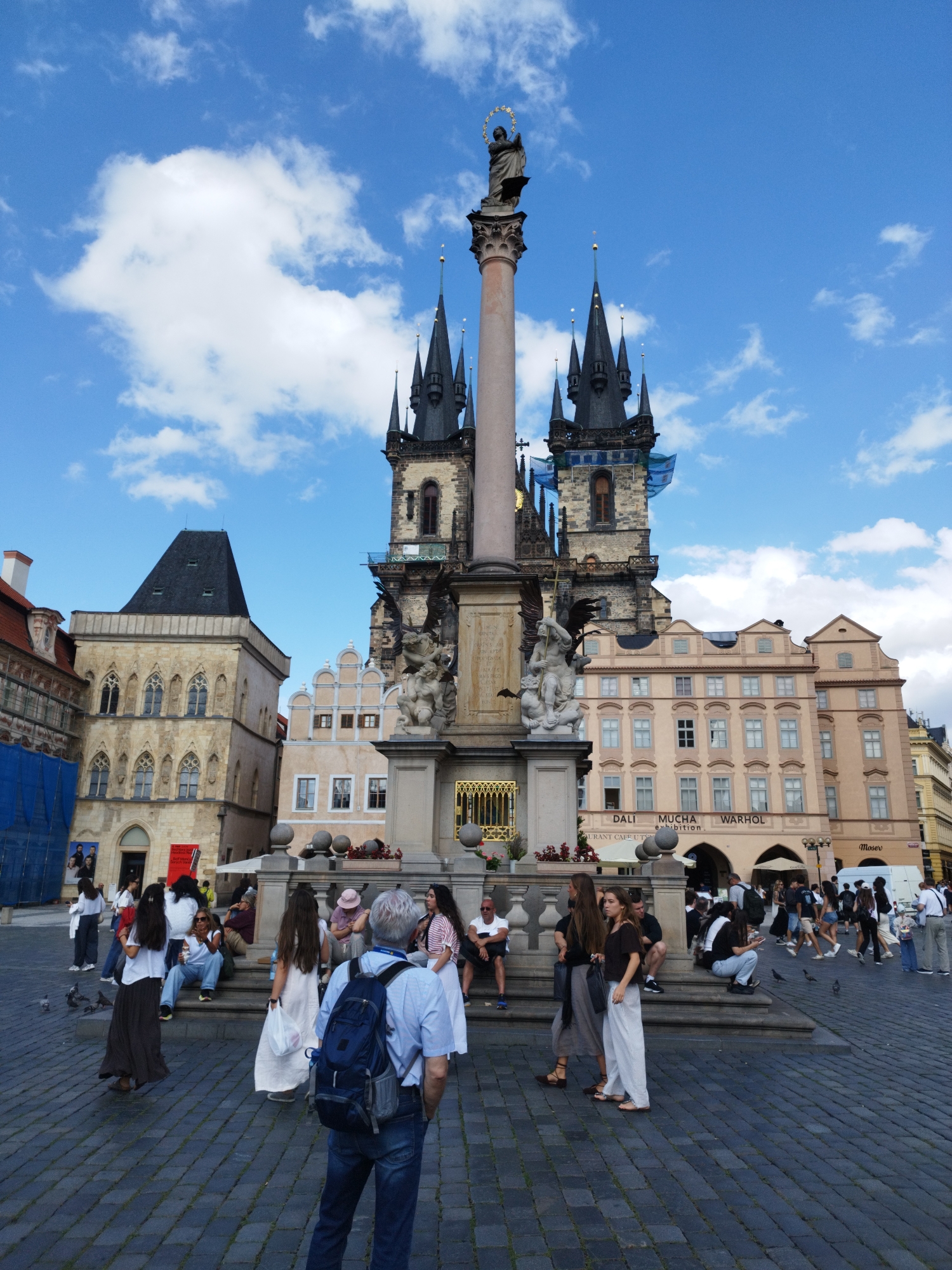 The image depicts a bustling public square with a central monument and surrounding buildings. The monument is a tall column topped with a statue of a figure with a halo. At the base of the column, there are sculptures of winged figures and possibly mythological creatures. Behind the monument, a gothic-style church with spires dominates the background. Several people are visible in the square, some standing, others sitting on the steps of the monument or on ledges. They appear to be tourists, dressed in casual attire. The square is paved with cobblestones. The sky is blue with scattered white clouds. The architecture of the buildings suggests a European city, likely with historical significance. One building has signage that includes "DALI MUCHA WARHOL exhibition."