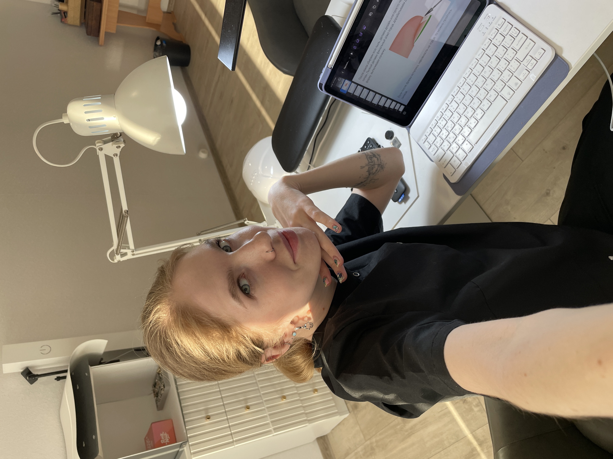 A selfie shot of a young woman with strawberry blonde hair and fair skin. She is wearing a black shirt and has a tattoo on her left forearm, visible as her arm rests on a white desk. Her nails are decorated with colorful designs. She has a nose ring and wears dangling earrings.
The scene appears to be indoors, possibly her workspace. A white adjustable desk lamp is positioned to her left, providing light. In front of her is a tablet with a white keyboard. The tablet screen displays a page with text in Russian. The background includes a white cabinet, a chair, and glimpses of a wooden structure.
The OCR text visible on the tablet seems to discuss nail care and shaping, suggesting she might be involved in the beauty industry.