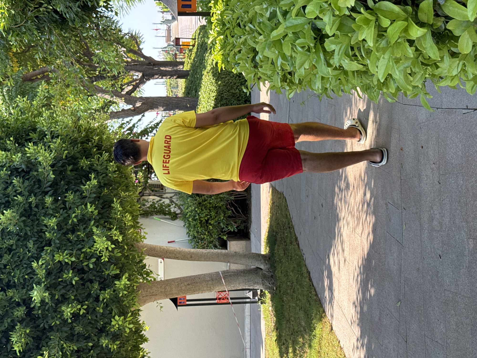 A lifeguard wearing a yellow shirt with "LIFEGUARD" printed on the back in red and red shorts walks away from the camera on a paved walkway. He is wearing white and black slides. To the left of the walkway is a patch of grass and a tree with a white building behind it. A red and white striped rope is tied around the tree. To the right of the walkway is a dense green bush. In the background, there are more trees and a playground area with structures. The scene appears to be outdoors in a sunny environment, possibly a resort or park in Aksu, Turkey.