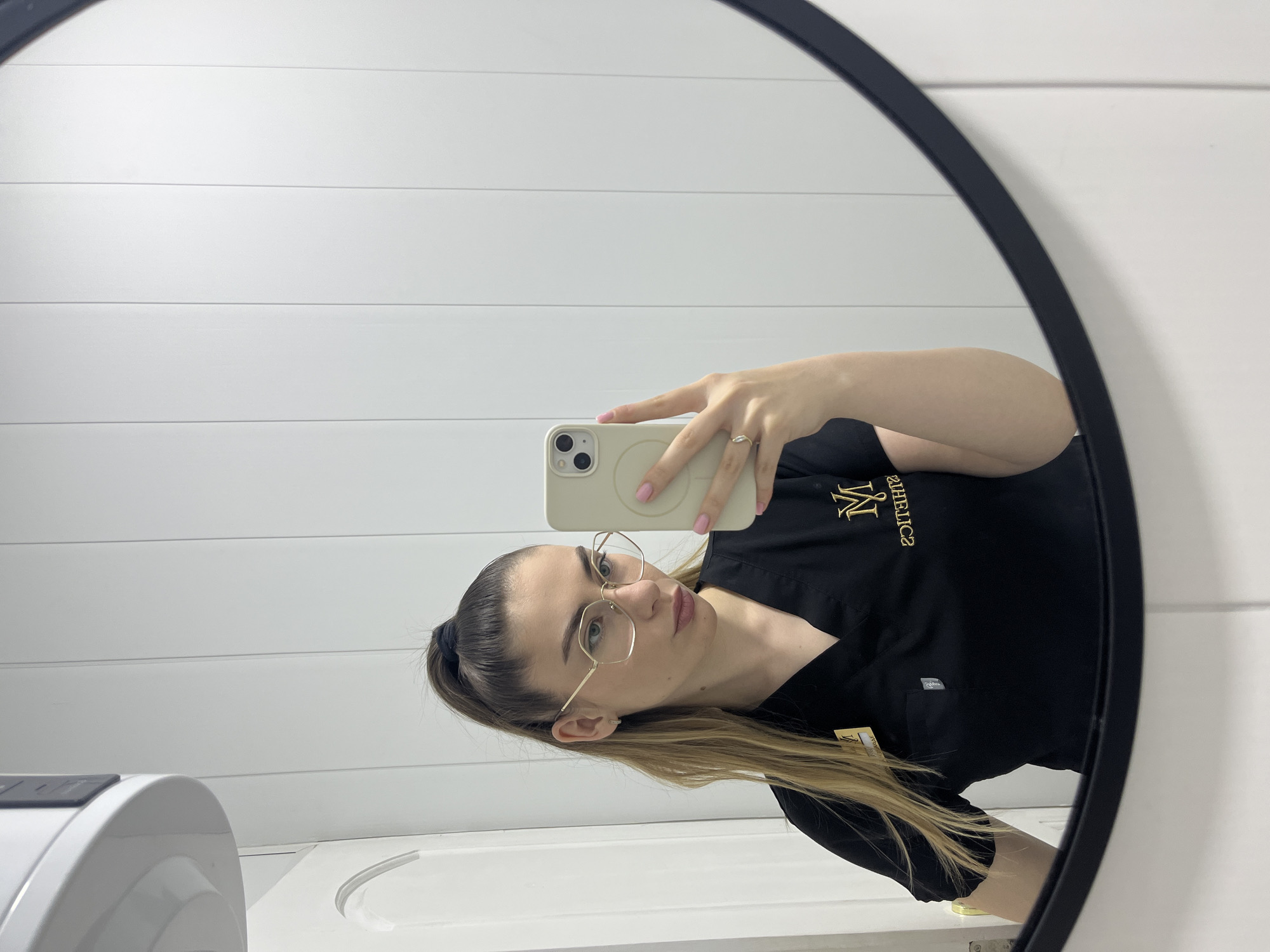 A woman with fair skin and long, blonde-streaked hair pulled back in a ponytail is taking a selfie in a circular mirror. She is wearing gold-framed glasses and a black uniform. The uniform has gold lettering on the left chest. She is holding a light-colored iPhone in her right hand. Her nails are painted pink. The background consists of white, horizontally-paneled walls. A white object is visible in the lower-left corner of the image. The city and exact location are unknown.