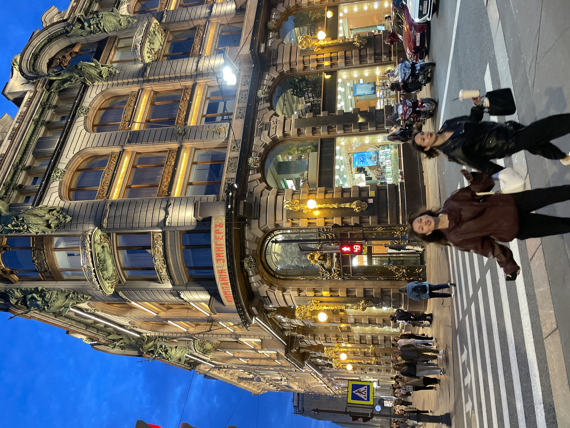 An eye-level, vertical, medium outdoor shot captures a bustling street scene in front of an ornate, lit building with significant architectural detail, possibly in a European city. The building features multiple stories with arched windows, decorative sculptures, and elaborate golden lampposts. A prominent red sign with Cyrillic text, "КОМПАНИЯ ЗИНГЕРЪ," is visible on the upper facade. The sky is a deep twilight blue.
In the foreground, a pedestrian crosswalk is occupied by several individuals. Two young women are walking across the street from left to right, with the woman on the right holding a drink and a handbag, and the woman on the left carrying a white bag. Behind them, a group of people walk towards the viewer. To the left, a pedestrian crossing sign and a traffic light showing red are visible.
In the mid-ground, several motorcycles and cars are stopped at the intersection. A red car and a white car are parked on the right side of the street. The building's ground floor appears to be a commercial space with large glass windows displaying merchandise and advertisements. The overall atmosphere suggests a vibrant urban environment at dusk.