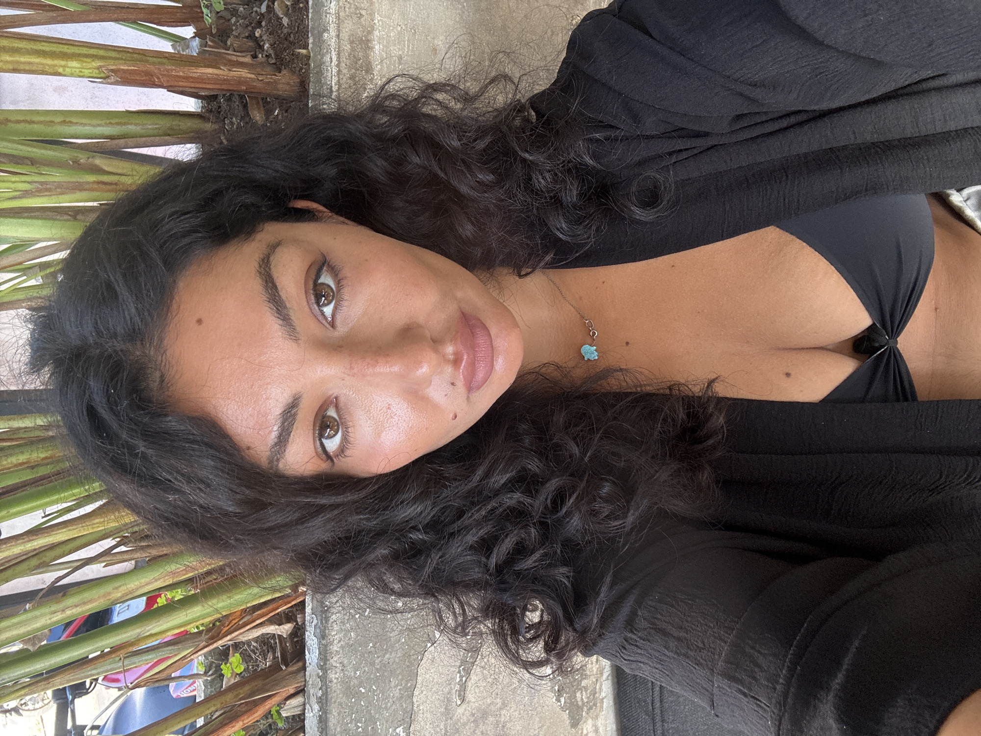 A woman with dark, curly hair, light brown eyes, and fair skin is seen in a selfie. She wears a black top with a low neckline and a sheer black cardigan. A turquoise necklace is visible around her neck. The background includes tropical vegetation and a concrete surface. The location is Kuta Selatan, Indonesia.
