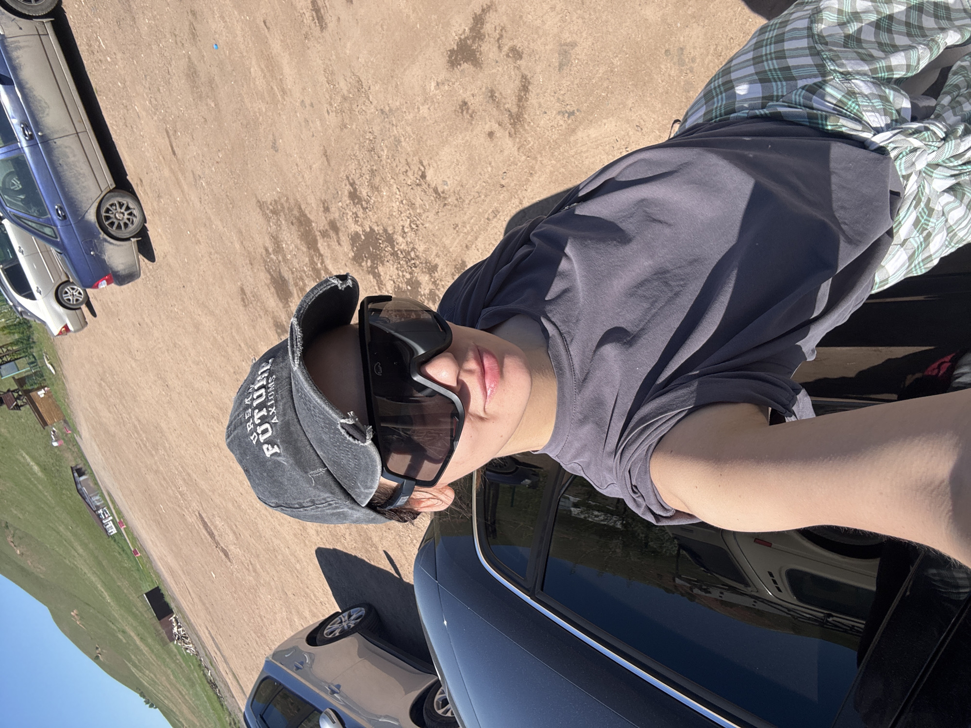 A young person is the central subject of this outdoor, brightly lit image. They are seen from the chest up, wearing a dark grey t-shirt, a distressed grey baseball cap, and large, dark sports-style sunglasses. A green and white plaid shirt is tied around their waist. The text "BREAK FUTURE AXIONS" is visible on the baseball cap. Their face, partially obscured by the sunglasses, shows neutral lips with a hint of pink lip gloss.
The setting appears to be an unpaved area, likely a dirt or gravel parking lot, under clear, sunny skies, evidenced by strong shadows. In the foreground, part of a dark-colored car (possibly black or dark blue) is visible, reflecting the environment. Further back, several other vehicles are parked, including a blue sedan and a white car. The left side of the background reveals expansive green grassy hills and distant structures, suggesting a rural or open landscape. The image is taken with a tilted perspective. Based on the context, the location is Дрокино, Russia.