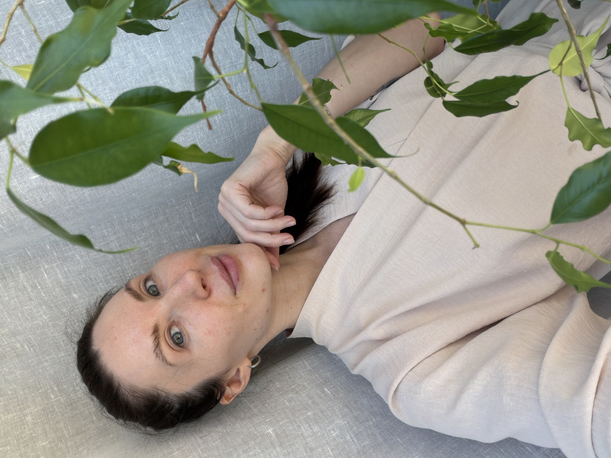 A woman with fair skin and dark hair is lying down, her head resting on a textured gray surface, likely fabric. She is wearing a light beige garment. Her hand is positioned near her neck. Green leaves and brown branches from a plant frame the upper part of the image, partially obscuring the scene. A location cannot be determined from visual information.