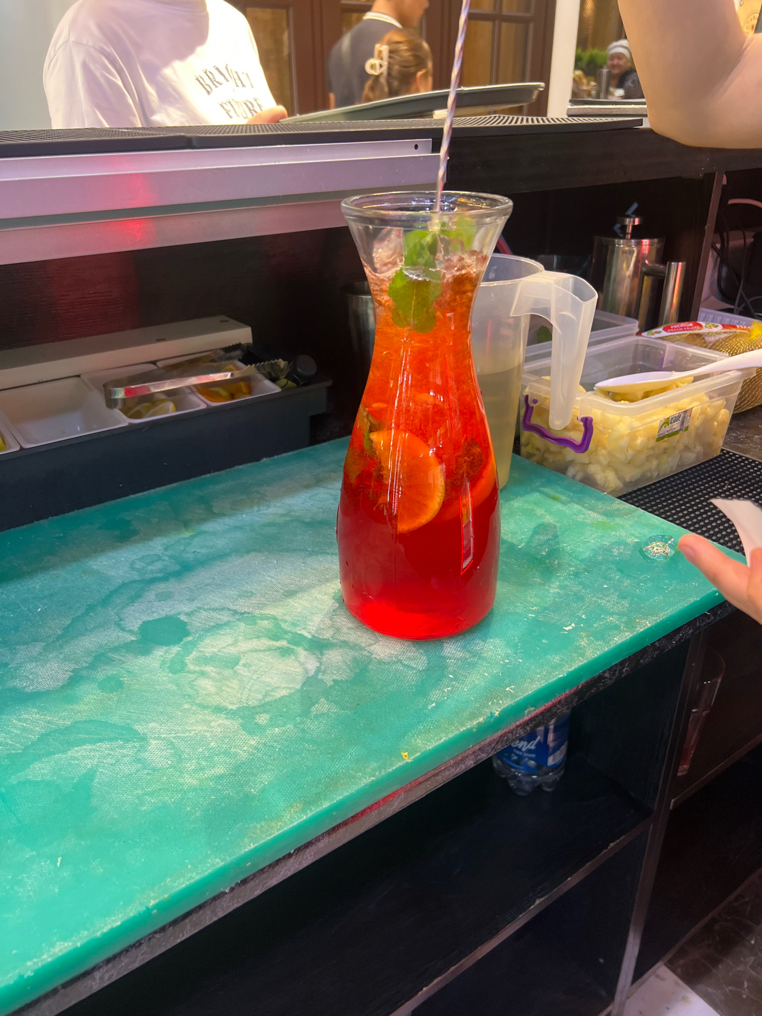 The image shows a close-up view of a bar area. In the foreground, a large glass pitcher filled with a red beverage, ice, mint, and orange slices sits on a turquoise cutting board. Behind the pitcher, there are several containers, one of which is filled with chopped fruit, likely pineapple, and another appears to be a water pitcher. A metal rod with a striped straw extends into the pitcher. In the background, a person wearing a white t-shirt with text printed on it is visible, as well as another person's arm and shoulder. A reflection in the mirror behind the bar shows a person wearing a white head covering. A blue plastic bottle is visible on a shelf below the cutting board. The overall environment suggests a food service establishment, possibly a cafe or restaurant. The location is specified as Osh City, Kyrgyzstan.