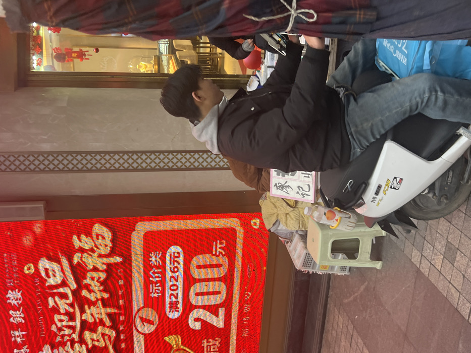 This image depicts an urban street scene in Jiangjin, China, in front of a commercial establishment. A male individual, wearing a black jacket, grey hooded sweatshirt, and blue jeans, is seated stationary on a white electric scooter. He is positioned facing slightly right, with his hands on the scooter's handlebars.
To the left, a large, vibrant red LED display or banner is visible, advertising "鼎祥銀樓" (Dingxiang Silver Building) with "HAPPY NEW YEAR 迎元旦 马年纳福" (Happy New Year, welcoming New Year's Day, blessings for the Year of the Horse) text. A promotional offer of "满2026元 减200元" (Spend 2026 yuan, get 200 yuan off) is also displayed in red and yellow text.
To the right of the LED display, a building facade with a light-colored wall features a vertical decorative grid pattern. A glass entrance door, adorned with red festive decorations, leads into a well-lit store, likely the "Dingxiang Silver Building," where display cases with gold-colored jewelry are visible, along with beige seating.
In the foreground, adjacent to the scooter, a light green plastic stool is present, featuring a bottle with orange liquid held in a yellow cup holder. A white sign with black Chinese characters, "廖记" (Liao Ji), rests near the scooter. A blue bag is placed on the ground next to the scooter's right side. Partially visible on the far right is a red and blue plaid fabric, possibly part of an awning or garment. The ground is paved with interlocking reddish-brown and grey tiles.