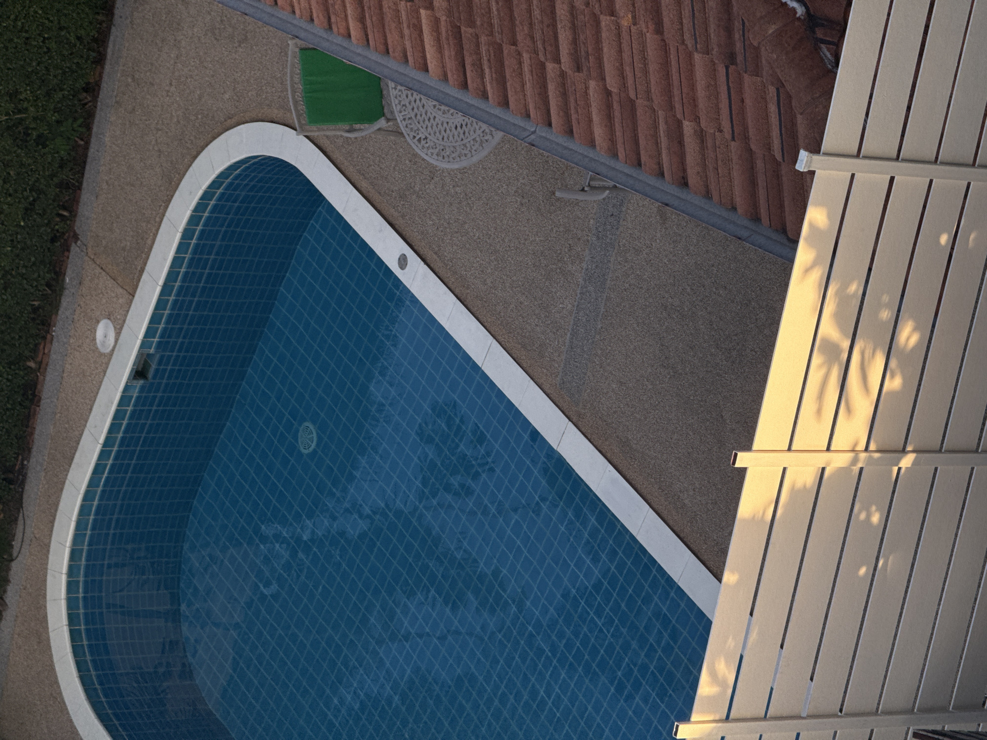 An aerial view captures a blue swimming pool with tiled walls and a white border, surrounded by a light brown speckled concrete deck. To the right of the pool, a red tiled roof of a building is visible, with a portion of its gray gutter system running along the edge. In the foreground, a horizontal light beige fence casts shadows of palm fronds onto its surface, suggesting the presence of nearby trees and the time of day. Adjacent to the pool and deck, a portion of dense green foliage forms a natural boundary. A round, white wrought-iron table with a matching chair, featuring green upholstery, is positioned on the deck near the pool's edge. The scene is set in Rawai, Thailand, a coastal district known for its beaches and resorts.