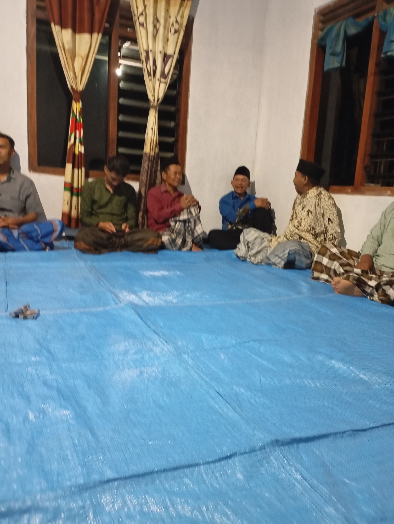 The image depicts a gathering of five men sitting on a blue tarp on the floor. The scene appears to be indoors, possibly in a room in Ketepung, Indonesia, given the provided context.
From left to right:
The first man is wearing a grey shirt and a sarong. He is looking towards the left.
The second man is wearing a dark green shirt and a sarong. He is looking down and appears to be holding something in his hands.
The third man is wearing a red shirt and a sarong. He is looking towards his right and seems to be engaged in conversation.
The fourth man is wearing a blue shirt and a sarong. He is smiling and looking towards his left.
The fifth man is wearing a patterned shirt (possibly batik) and a sarong. He is looking towards his left and appears to be in conversation with the man in the blue shirt.
The men are all seated cross-legged. The environment includes windows with patterned curtains and what appears to be a simple interior wall. A pair of sandals is visible on the blue tarp in the foreground. The lighting suggests it is evening or indoors with artificial lighting.