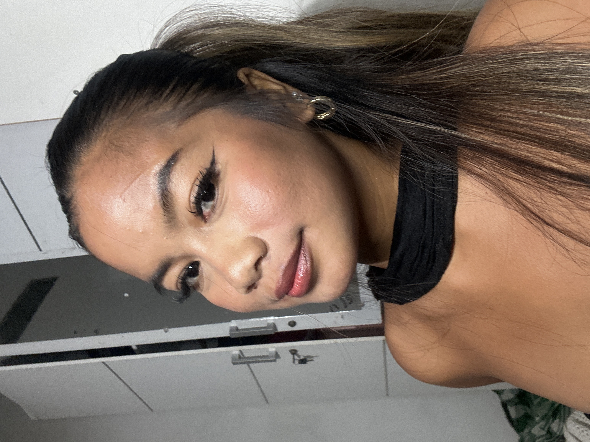 A close-up shot of a young woman with light skin, wearing makeup that includes winged eyeliner and lip color. Her hair is styled in a partial ponytail, with brown hair at the front and black hair near the roots. She is wearing a black top with a high neckline. A gold hoop earring is visible in her left ear. The background shows what appears to be a white cabinet with silver handles. The location is potentially North Jakarta, Indonesia, but the image doesn't provide definitive evidence of the exact city.