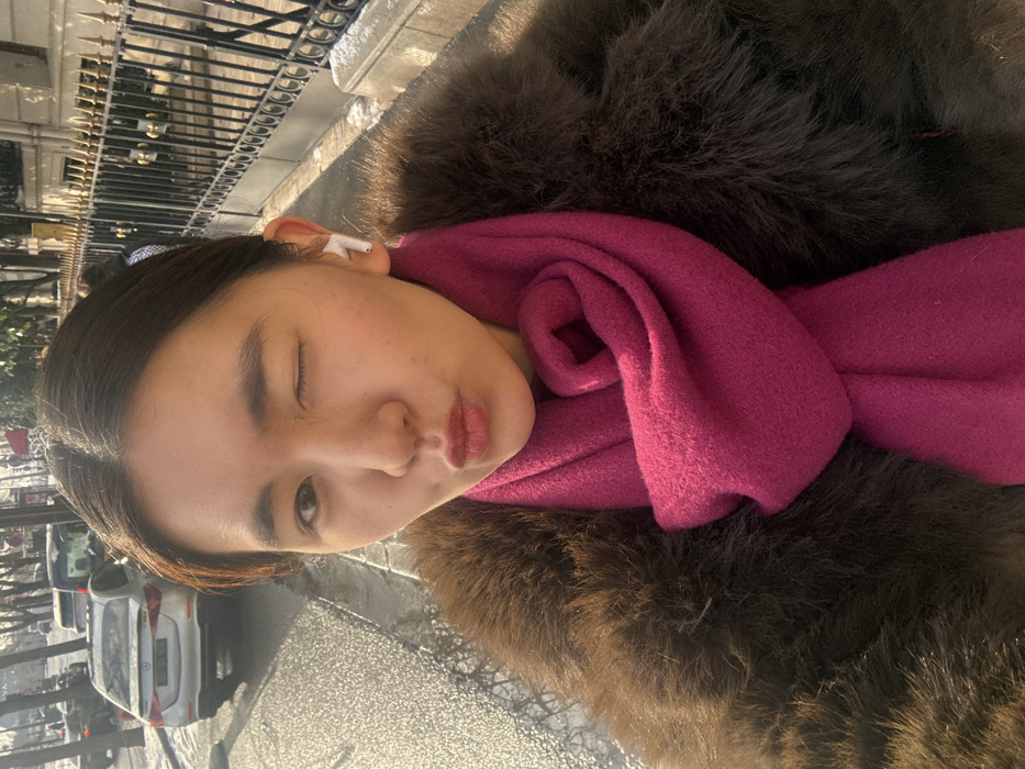 A medium-close-up selfie captures an East Asian female on a Parisian street during daylight. She winks with her right eye and pouts her lips, looking directly at the camera. Her dark hair is pulled back, with a small bun visible above her right ear. A white wireless earbud is present in her right ear. She wears a brown fur or faux fur coat and a bright fuchsia-colored scarf wrapped around her neck. The background features a wet asphalt street reflecting sunlight, indicative of recent precipitation or melting snow. Patches of snow are visible on the sidewalk and along the curb. To the left, a silver Mercedes-Benz A-Class sedan is parked or moving, displaying a French license plate (partially visible "FR"). Further down the street, other vehicles and bare trees are visible. To the right, a classic Parisian building facade is partially obscured by a black ornamental iron fence with gold-colored decorative elements.
