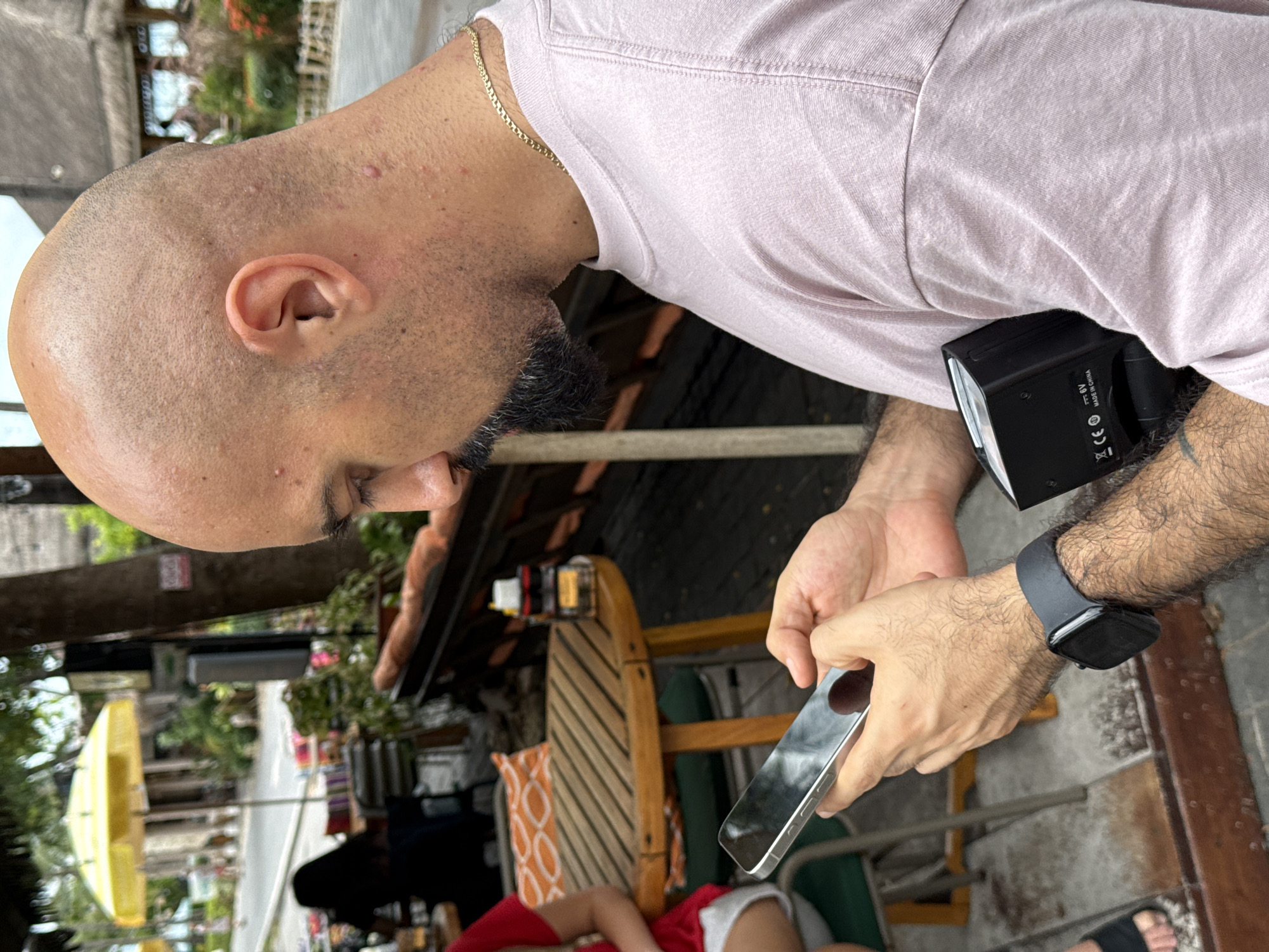 A man with a shaved head and a goatee is holding a smartphone in his hands, looking down at it. He is wearing a light pink t-shirt and a gold chain. On his left wrist, he has a black smartwatch. In his right hand, he is holding a black rectangular object that appears to be a camera flash. He is seated at an outdoor cafe with a wooden table and chairs. In the background, there is a yellow umbrella and some vegetation. The setting appears to be a street in Ko Samui District, Thailand.