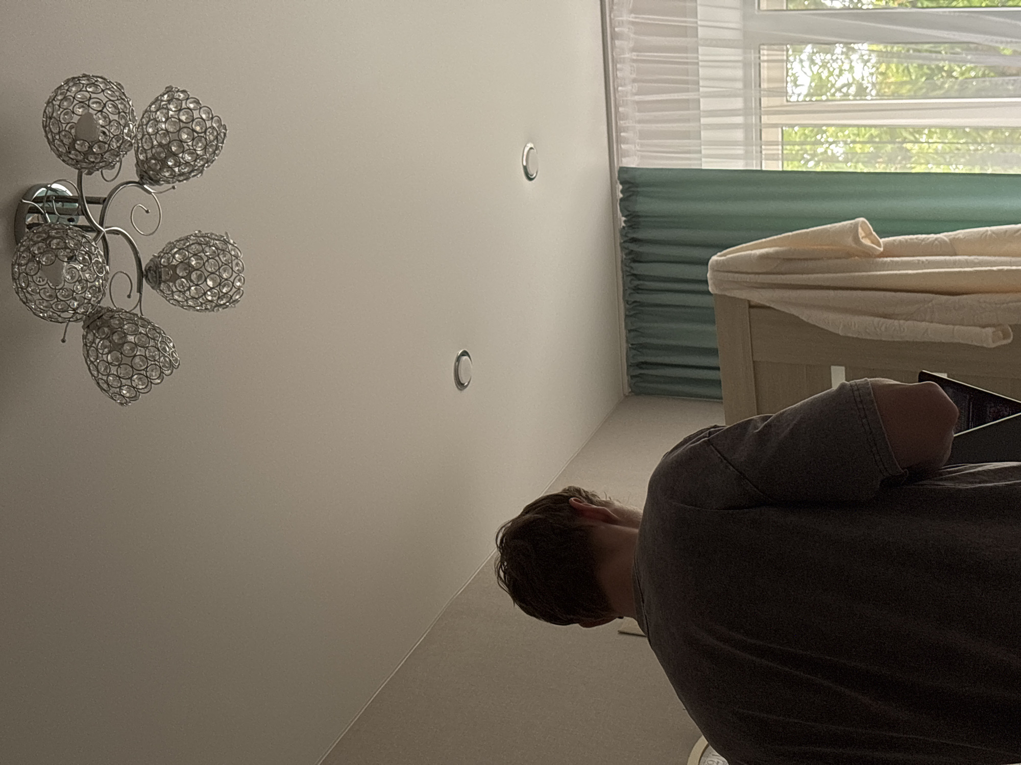 The image shows the interior of a room, likely a bedroom. The perspective is from behind a person facing away from the camera, wearing a dark t-shirt. The person's back and the side of their head are visible. In the upper part of the image, a chandelier with multiple crystal-like globes is attached to the ceiling. Two circular ceiling lights are also visible. The walls are light brown and a light-colored curtain is visible in the background, with a translucent white curtain over a window to the right, through which trees and sunlight are visible. A towel or robe hangs on a piece of furniture on the left side of the image. The overall impression is of a domestic interior. There is no specific identifiable information to place this scene within the city of Sarov, Russia.