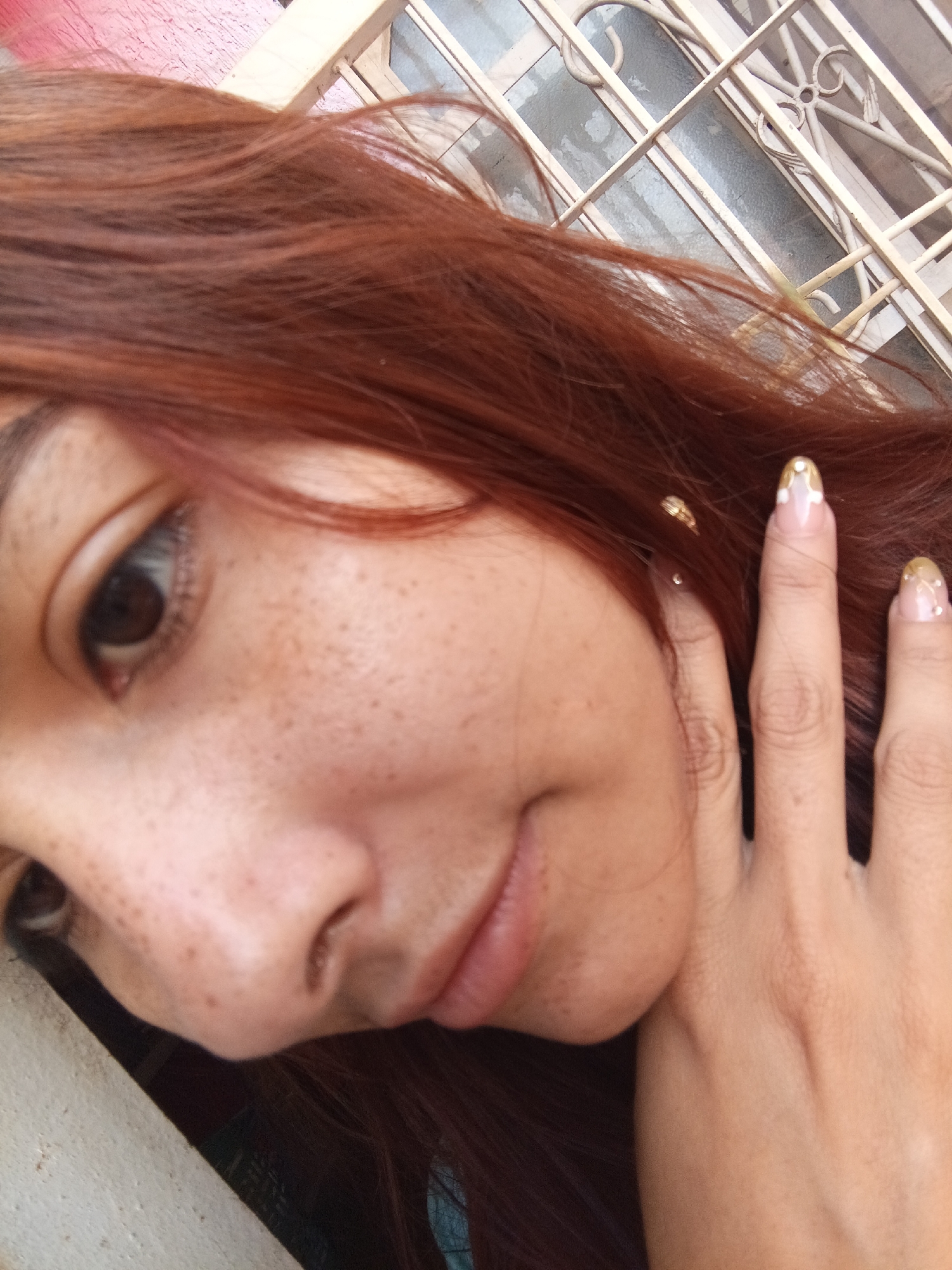 A close-up selfie shows a person with reddish-brown hair, light skin covered in freckles, and a hand with decorated nails. The nails have a clear base with gold tips and white floral accents. The background features a white metal grate, possibly a window or gate, and hints of a painted red wall. The location is unknown, and it's impossible to determine the exact location is Juan Griego, Venezuela, based on the information provided in the image.