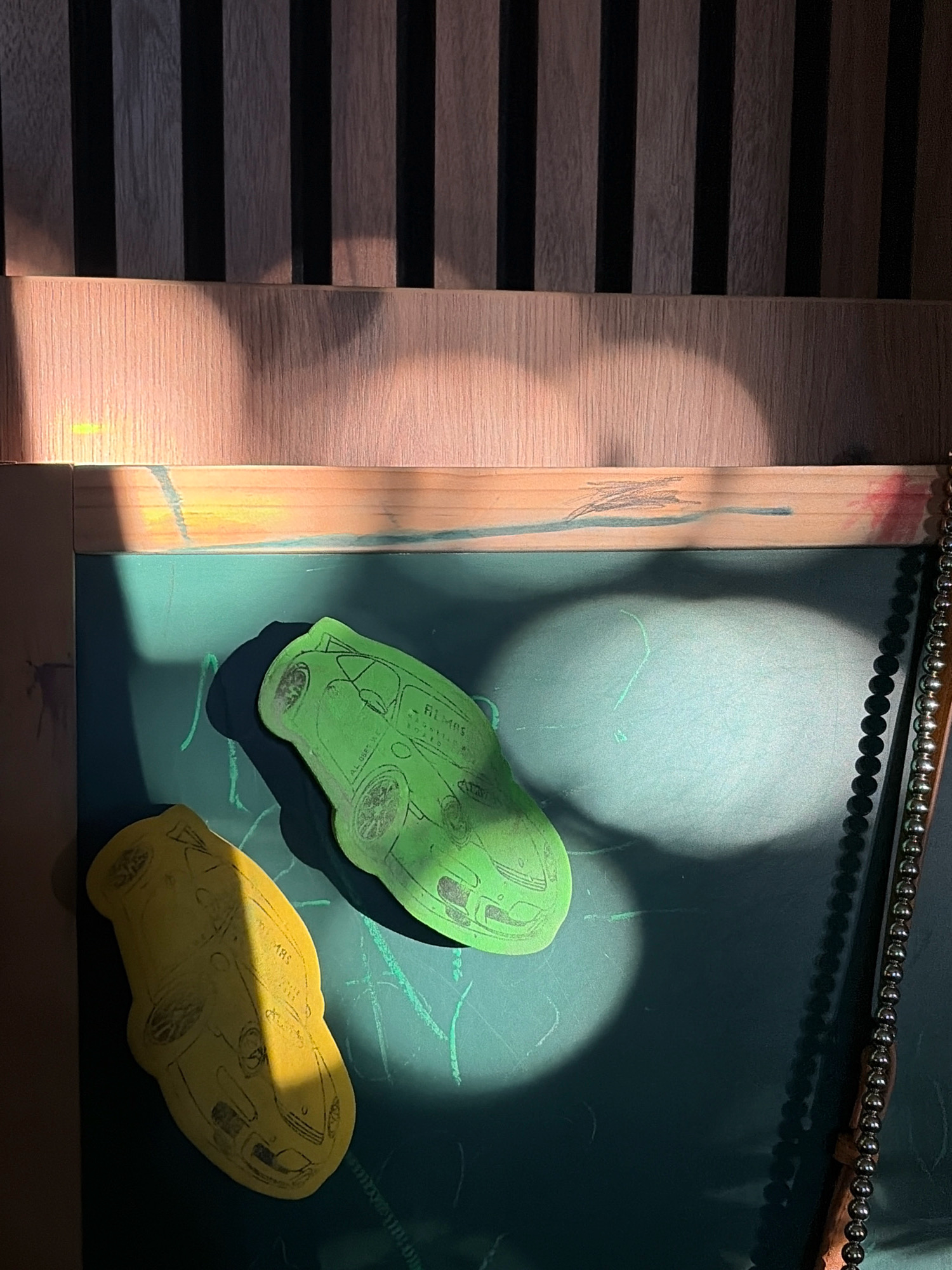 The image depicts a close-up view of a dark green chalkboard on a wooden easel, illuminated by natural light casting strong shadows. Two car-shaped cutouts, one green and one yellow, are affixed to the chalkboard. The green cutout features an illustration of a van with the text "ALMOS" and other illegible writing. The yellow cutout shows a drawing of a vintage Volkswagen Beetle with the text "ABS". Green chalk marks are visible on the chalkboard's surface, suggesting it has been used for drawing or writing. The top of the easel is framed by a wooden border with faint colorful markings. A beaded decorative edge runs along the right side of the easel. The background consists of vertical wooden slats, creating a striped pattern. The scene appears to be indoors, given the presence of the chalkboard and easel. There are no people or animals in the image. The overall impression is of a creative or educational setting, possibly a home or classroom. No information regarding Oxentea, Moldova is discernible from the image.