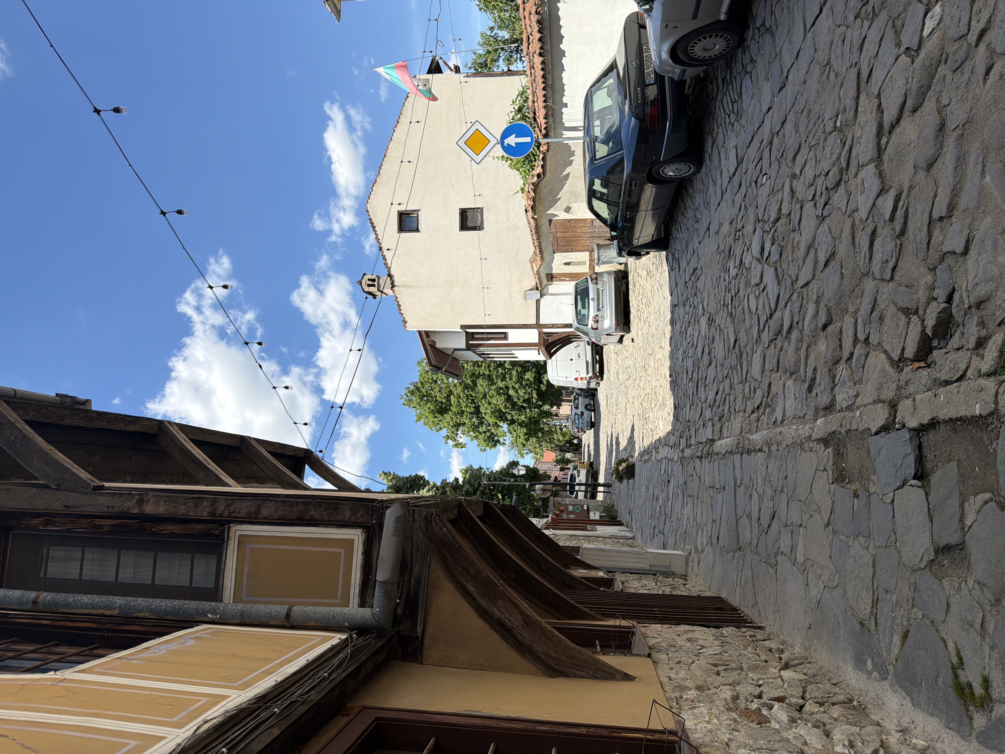 The image depicts a narrow street in the Old Town of Plovdiv, Bulgaria. The street is paved with cobblestones and slopes upwards. On the right side, a dark-colored Mercedes-Benz car is parked. Further down the street, other parked vehicles are visible. A building with a cream-colored facade and a red-tiled roof stands on the right. A Bulgarian flag is attached to the top of the building. A blue and white road sign with a left arrow and a diamond-shaped sign are mounted on a pole. On the left side of the street, the edge of a building with wooden structural elements and windows is visible. Overhead, a string of decorative lights spans across the street. The sky is blue with scattered white clouds. The lighting suggests it is daytime.