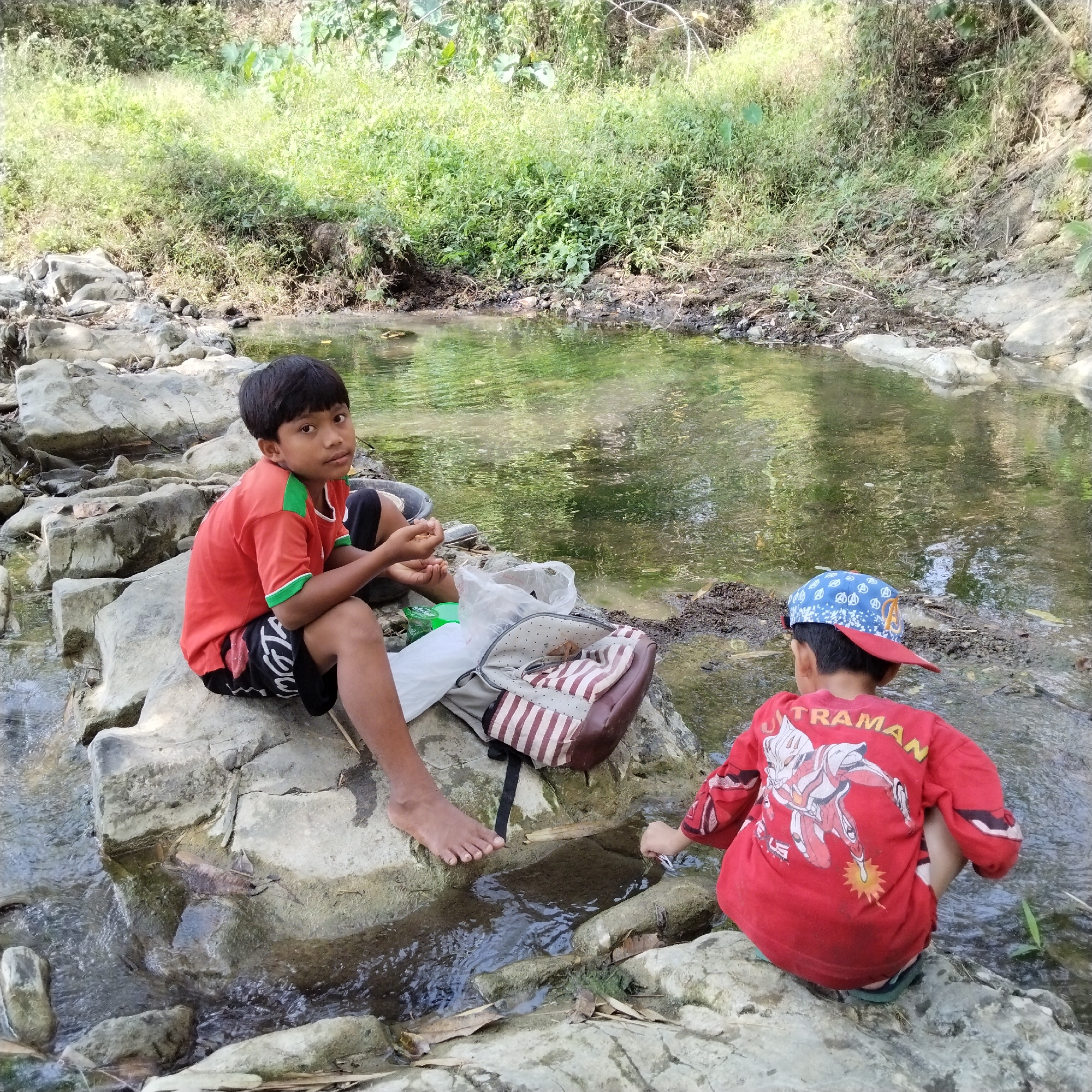 In Karangbaru, Indonesia, two young boys are by a shallow stream with rocky banks. The boy on the left, wearing a red t-shirt with green trim and black shorts, sits on a large rock facing forward, looking at the camera. A striped backpack lies beside him. The boy on the right, wearing a red t-shirt with an Ultraman graphic and a blue cap, is crouched by the water, his back to the camera, interacting with the stream. The stream is clear, reflecting the lush green vegetation that lines the opposite bank.