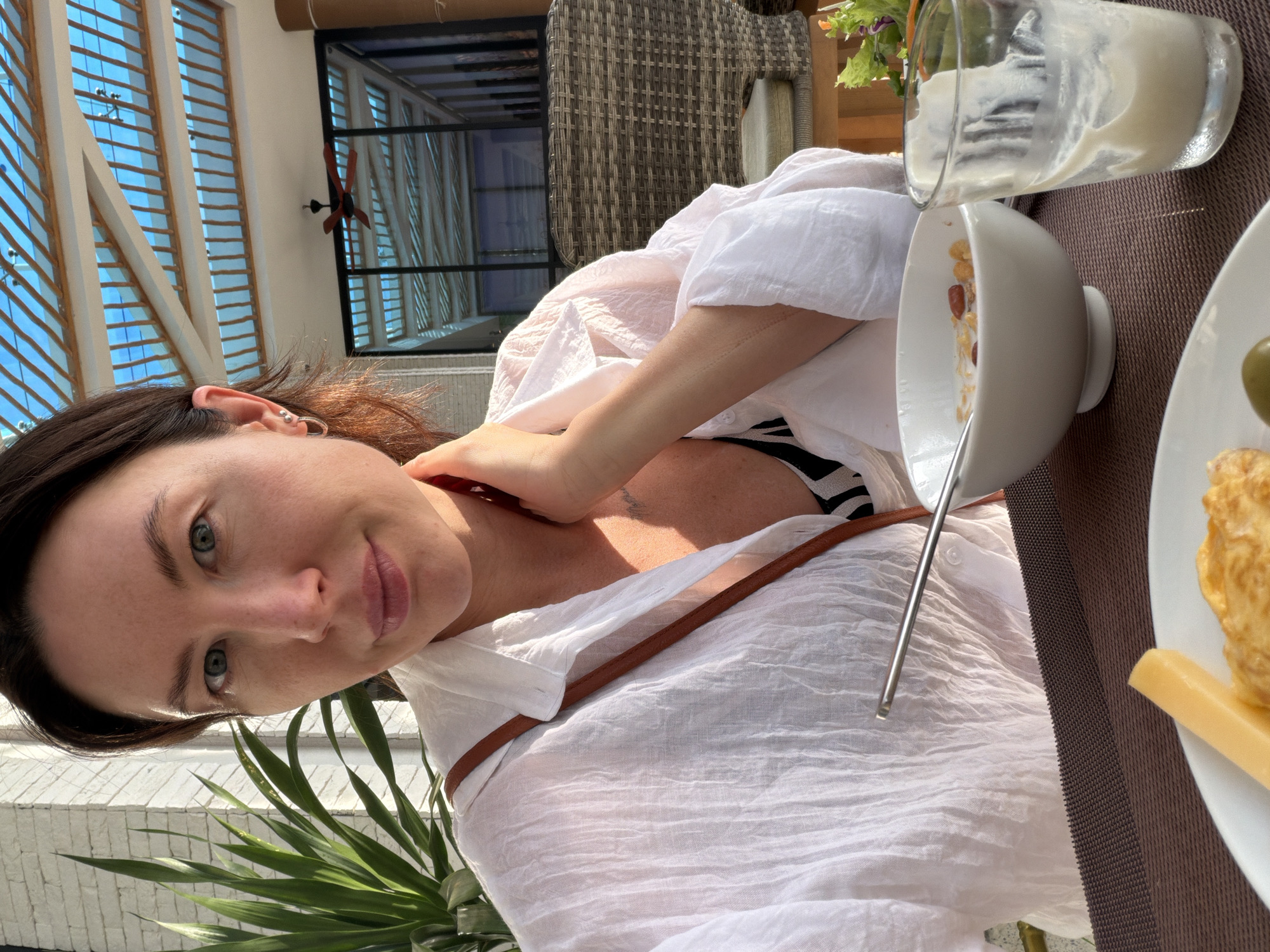 A woman with dark hair is seated at an outdoor table, taking a selfie. She is wearing a white button-down shirt over a black and white patterned top. Her right hand is touching her neck, and a brown strap is visible across her shoulder. In front of her are a bowl of cereal, a glass with a white liquid, and a plate with cheese and an olive. Behind her, a ceiling fan is visible within an enclosed area with blue tinted windows. The scene appears to be in a restaurant or cafe with wicker chairs and a plant. The location is specified as Dong Ninh Hoa Ward, Vietnam, implying it's an establishment within that area.
