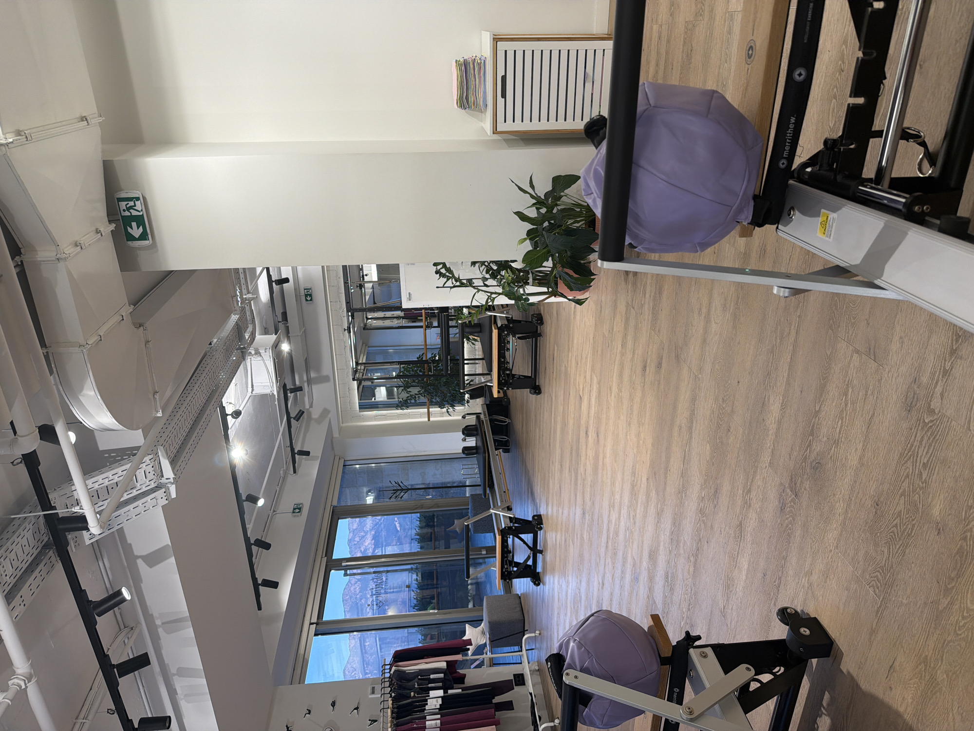 The image depicts the interior of a Pilates studio located in Boreti, Montenegro. The studio features a modern, minimalist design with light-colored wooden floors and white walls. Large windows offer a view of mountains and a cloudy sky, suggesting a daytime setting.
Several pieces of Pilates equipment are visible, including reformers and cadillacs, some of which are positioned in the foreground and mid-ground. Two large, light purple cushioned objects, possibly exercise balls or parts of equipment, are placed on some of the reformers. Clothing racks with athletic wear are set up near the windows on the left side of the frame.
In the background, there are more Pilates apparatus, plants in pots, and a white cabinet with a stack of folded towels on top. An illuminated green exit sign with an arrow pointing left is affixed to the ceiling near a pillar. The lighting in the studio appears to be a mix of natural light from the windows and overhead artificial lighting. The scene is devoid of any people, indicating the space is currently unoccupied.