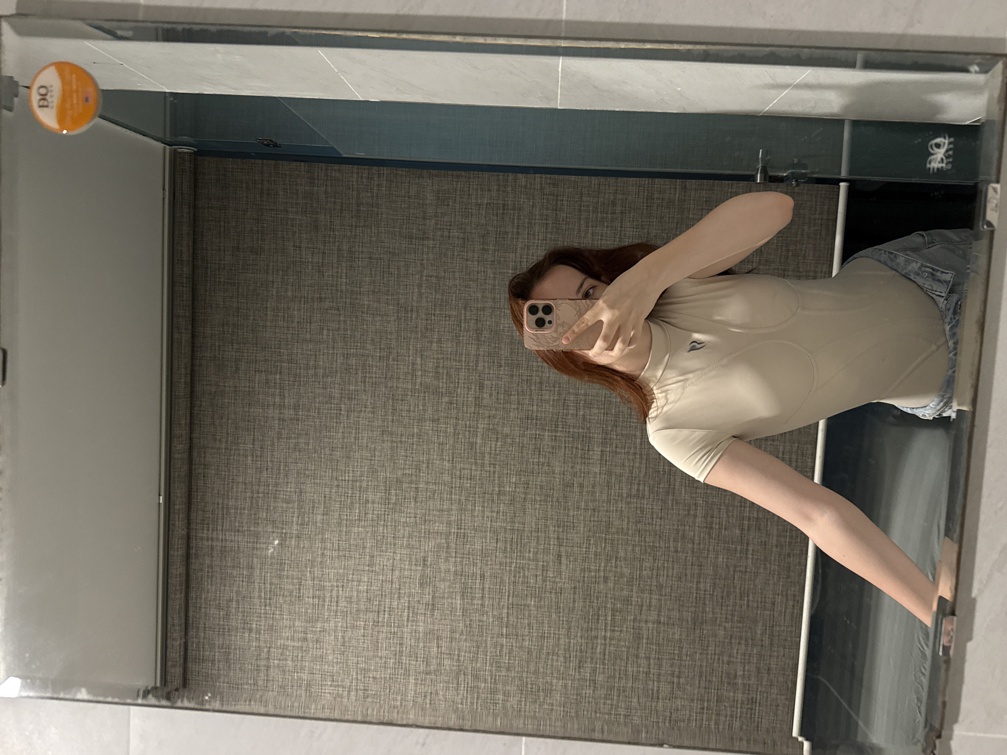 The image depicts a person taking a mirror selfie in what appears to be a bathroom. The person is holding a smartphone in their left hand, obscuring part of their face. They are wearing a beige-colored bodysuit and denim shorts. The background shows a textured wall and the edge of a shower or bathtub. A circular object with orange writing is visible on the mirror. It is unclear if the image was taken in the city of Bac Nha Trang Ward, Vietnam.
