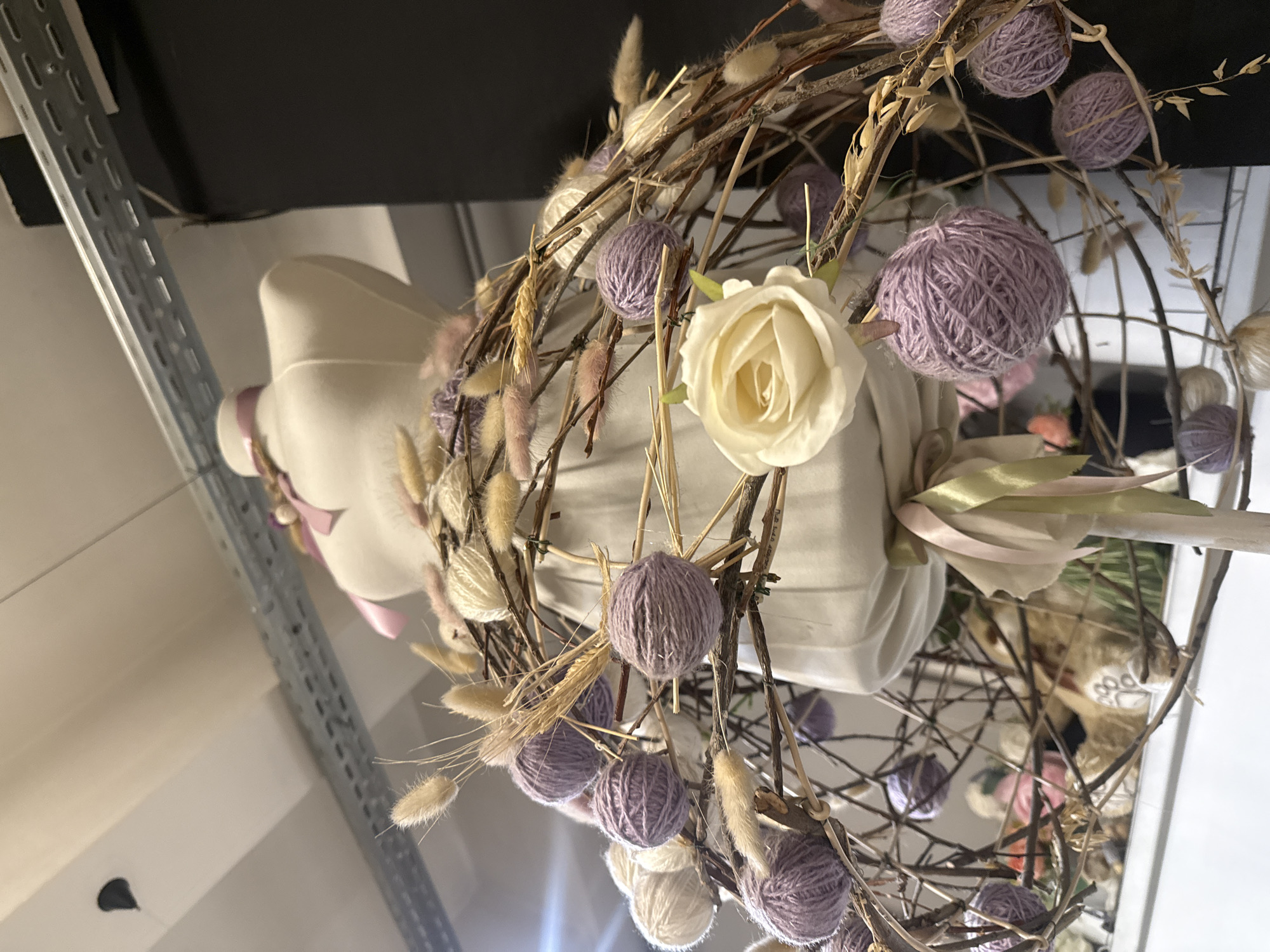 The image displays a decorative wreath or garland, oriented sideways as if viewed from below and to the side, within an indoor setting in Yekaterinburg, Russia. The primary subject is a rustic wreath constructed from intertwined dark brown branches, adorned with numerous small, spherical balls of yarn in varying shades of lavender and cream.
Interspersed among the yarn balls and branches are dried natural elements, including fluffy, light-colored seed heads resembling pampas grass or bunny tails, and stalks of dried wheat or oats. A prominent artificial white rose with tightly packed petals is centrally placed within the upper part of the wreath.
In the background, partially visible behind the wreath, is the upper torso of a light-colored mannequin, possibly a dress form, draped with soft fabric. Delicate pink and pale green ribbons are tied around the mannequin's neck or shoulder. To the far left, a silver-colored, perforated metal shelving unit or support structure extends diagonally, suggesting a commercial space, workshop, or storage area. The scene is illuminated by artificial indoor lighting. No visible text is present.