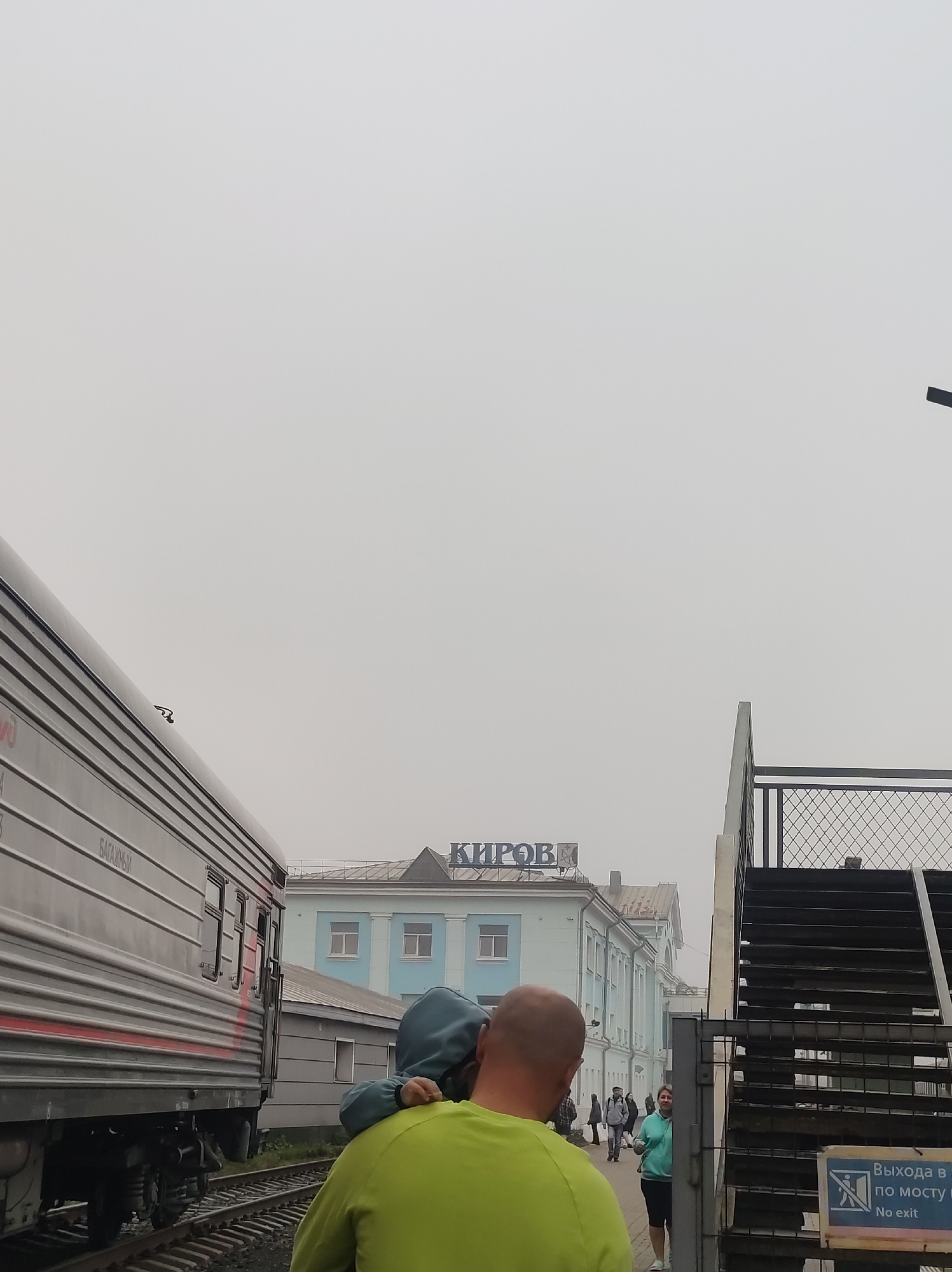 The image depicts a scene at a train station in Kirov, Russia. In the foreground, a person wearing a green shirt has another person, possibly a child wearing a blue hoodie, on their shoulders. To the left, a train car is visible, its ribbed metal siding a prominent feature. In the background, a light blue building with white trim and several windows stands behind a low gray structure. Above the building, a sign with the Russian word "КИРОВ" (Kirov) is visible. To the right, a metal staircase with diamond-shaped grating leads upwards, with a sign that says "Выхода в по мосту" and "No exit". Several other people are walking in the background near the station building. The sky is overcast and hazy, suggesting a cloudy or foggy day.