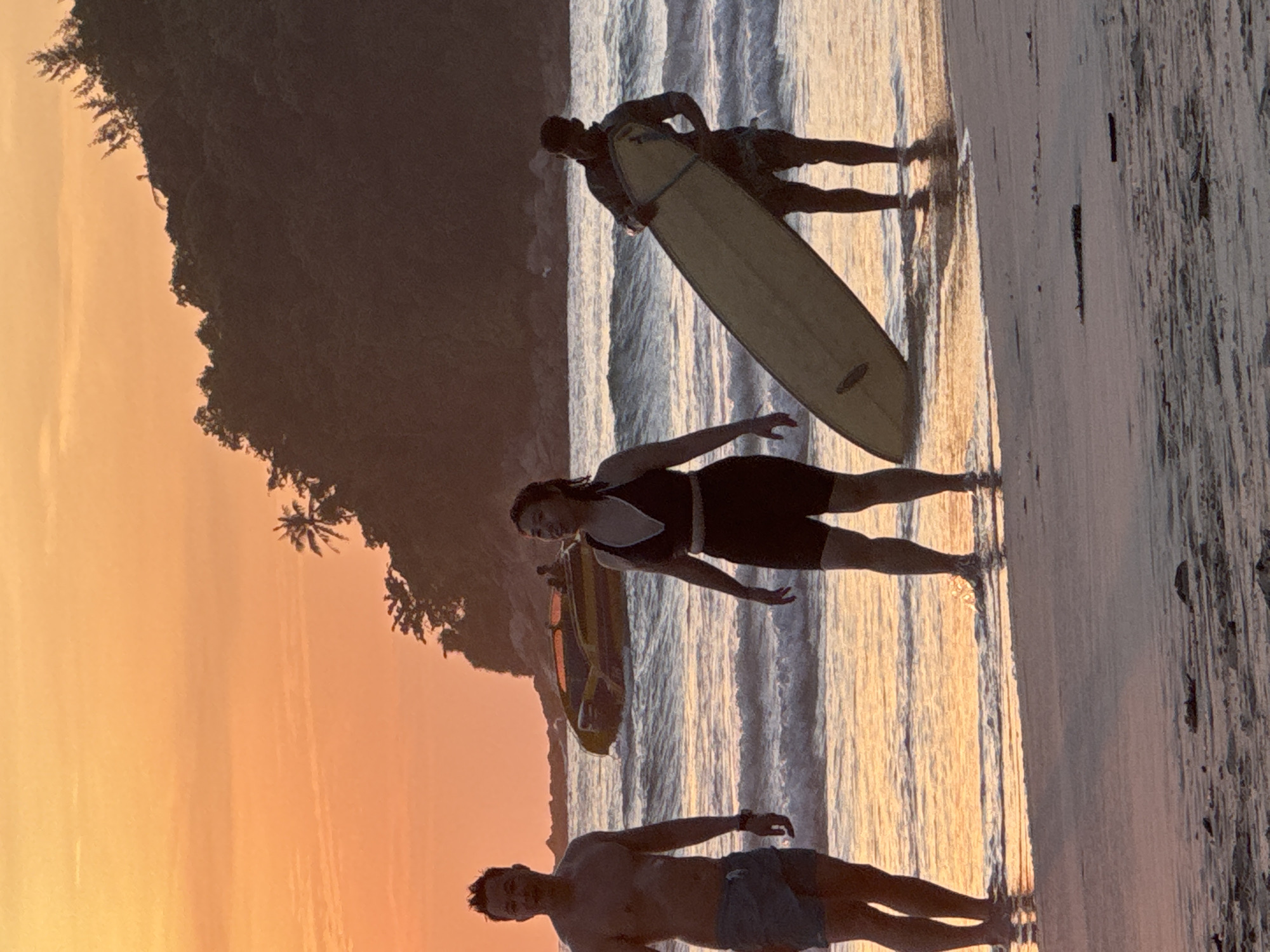 This image captures a serene sunset scene on Karon Beach, Thailand. The sky is awash in a vibrant orange hue, casting a warm glow over the tranquil sea. In the foreground, three individuals are enjoying the beach.
On the left, a man stands shirtless, wearing blue shorts, facing the camera with a slight smile. To his right, a woman in a black two-piece bikini stands facing away from the camera, her hands at her sides, her head turned slightly as if in conversation. Further to the right, another man is seen holding a surfboard vertically, partially submerged in the shallow water. He is wearing patterned shorts and a dark shirt, with his back to the camera.
In the background, a yellow and black speed boat is visible on the water, adding a touch of activity to the peaceful setting. The coastline features a lush, tree-covered hill, characteristic of Karon's landscape, silhouetted against the colorful sunset. Gentle waves lap at the shore, creating a soothing rhythm. The sandy beach is marked with scattered debris, hinting at the natural elements of the environment. The overall impression is one of a relaxed and picturesque evening on a tropical beach.