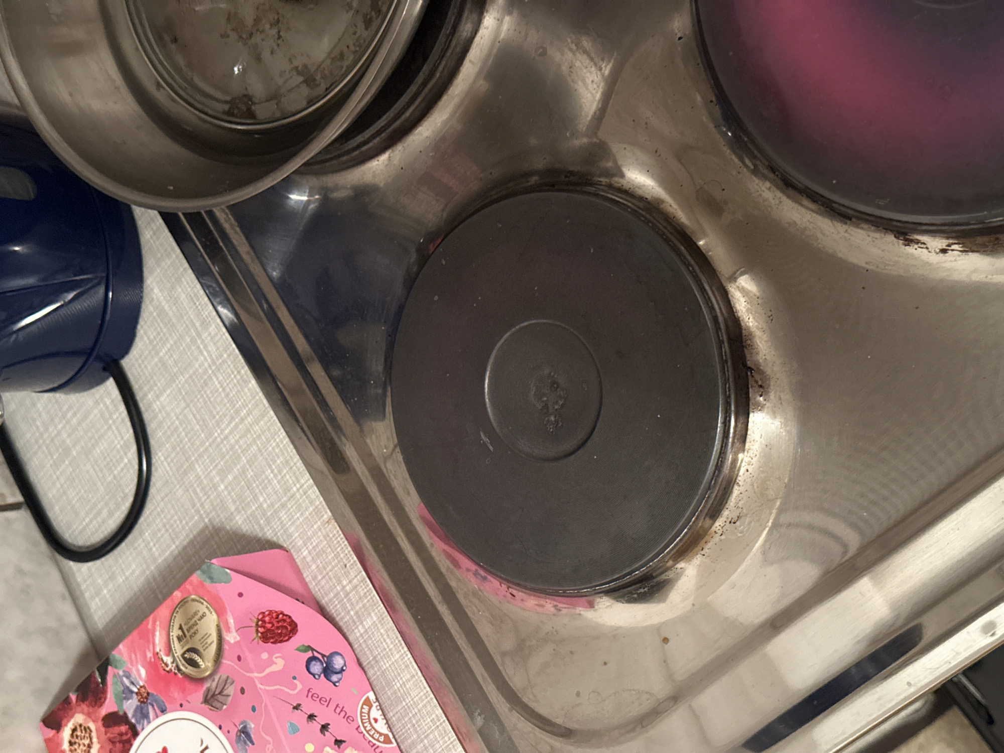 The image shows a close-up view of a four-burner electric stove with a stainless steel top. The burners are black and circular, with a visible heating element in the center of one. The stove is clean in some areas but shows signs of grease and food residue, particularly around the edges of the burners. To the left of the stove, partially in the frame, is a pink cardboard box decorated with floral patterns and images of berries. The box has text in a foreign language and a circular gold seal. In the background, the edge of a blue electric kettle is visible, along with a black electrical cord. The scene appears to be in a kitchen. There is no indication of the location being in Bischofswiesen, Germany.