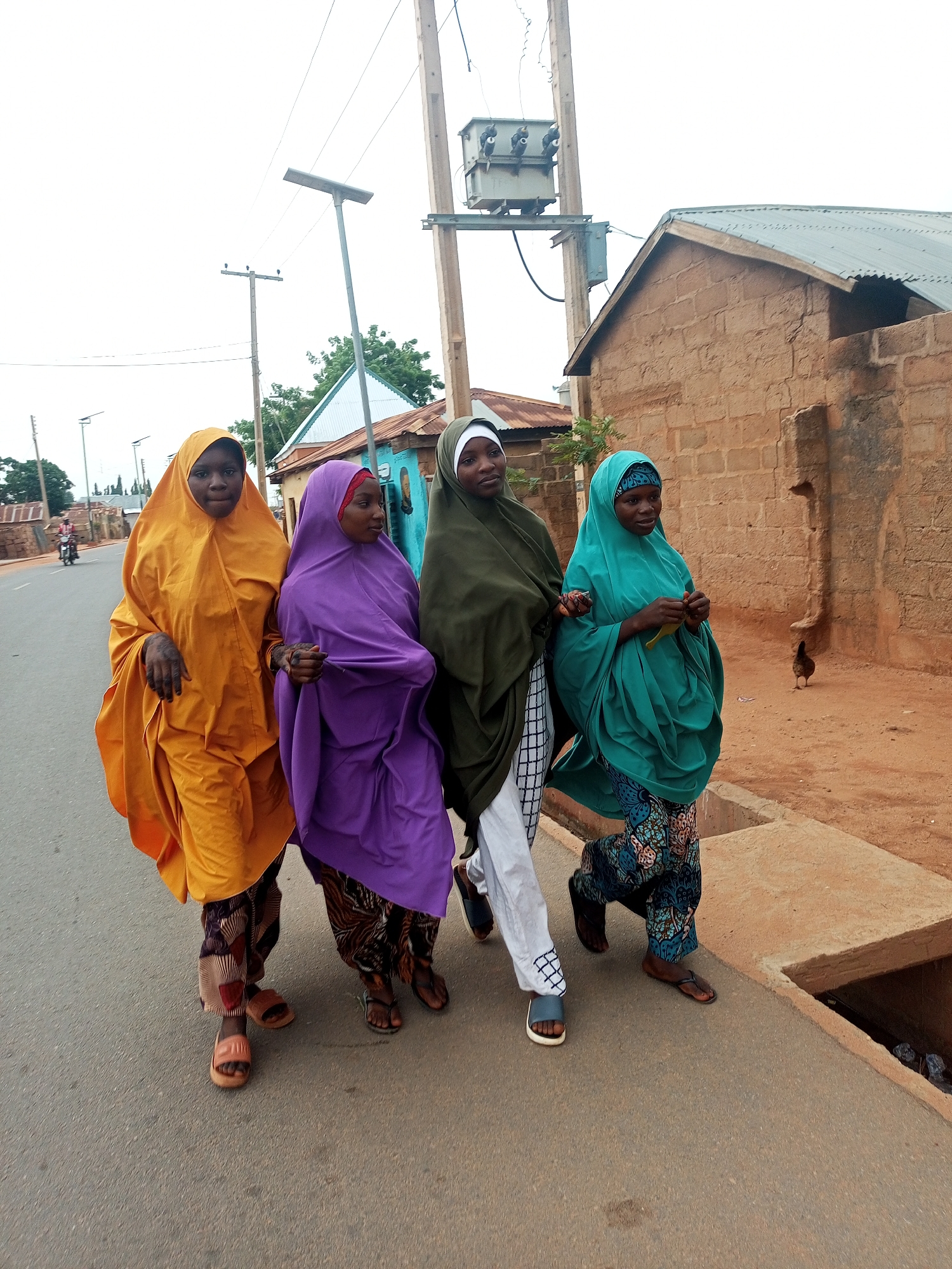 Four women wearing colorful hijabs are walking arm in arm down a paved street. The woman on the left is wearing an orange hijab, the second woman is wearing a purple hijab, the third woman is wearing a green hijab, and the fourth woman on the right is wearing a teal hijab. All four women are wearing long patterned pants and sandals. They are walking on the left side of the street, next to a curb. Behind them, there is a two-story brick building and a wooden utility pole with a transformer on top. In the background, there is a motorcyclist riding down the street. The location appears to be a residential area.