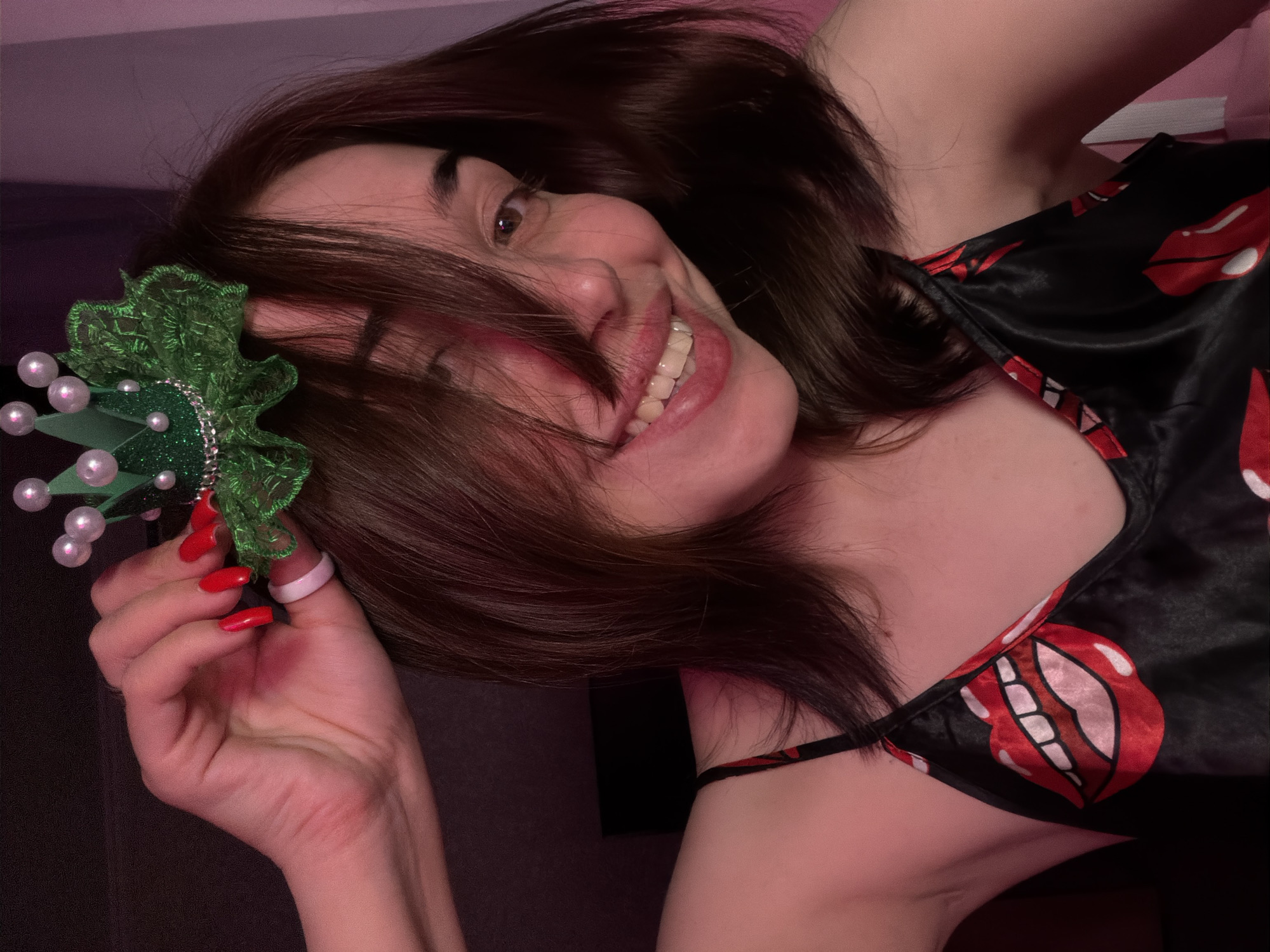 A young woman with brown hair and fair skin is shown in a close-up, head-and-shoulders shot. She is wearing a black slip with a pattern of red lips. She is also wearing a small, green, sparkly crown with pearl-like accents on her head. Her right hand is raised, holding the crown in place. Her left hand is visible around her left shoulder. The background is dark with pinkish lighting. The woman is smiling broadly, showing her teeth, and her eyes are looking towards the camera.