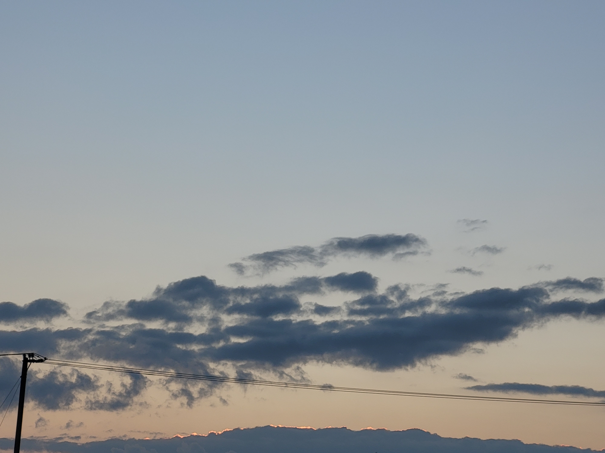 This is a photograph taken in the city of Ozolnieki, Latvia, featuring a sky filled with scattered clouds at sunset. A utility pole stands on the left side of the frame, with three wires stretching horizontally across the lower part of the image. The clouds are a mix of dark gray and lighter hues, with a thin band of orange light visible along the bottom edge, indicating the setting sun. The sky transitions from a pale blue at the top to a softer, warmer tone towards the horizon.