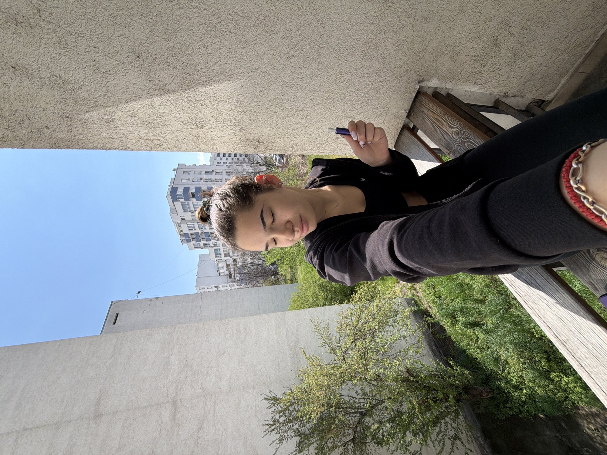 A young person, likely female, is captured in a bright outdoor selfie taken from a low angle. She has dark hair tied back in a bun and is wearing a black long-sleeved top or hoodie and black leggings. A bracelet featuring gold-colored chains and a red string is visible on her right wrist. In her left hand, which has white nails with black spotted accents, she holds a small, dark blue or purple cylindrical object, possibly a vape pen. Her eyes are closed or partially closed, and her head is tilted downwards, conveying a relaxed or contemplative mood.
The scene is set on what appears to be a balcony or terrace. Above the person, a textured, light-colored concrete or stucco ceiling casts a noticeable shadow. To the right, part of a wooden railing or bench is visible. The background reveals an urban landscape under a clear blue sky, indicative of a sunny day, likely midday or early afternoon. Several modern apartment buildings with multiple windows and balconies are scattered across the horizon. Below these buildings, lush green trees and grass suggest a landscaped area or park. A plain, light-colored concrete wall forms part of the immediate surroundings. The overall impression is a peaceful moment in an urban residential area in Sofia, Bulgaria. No visible text is present.
