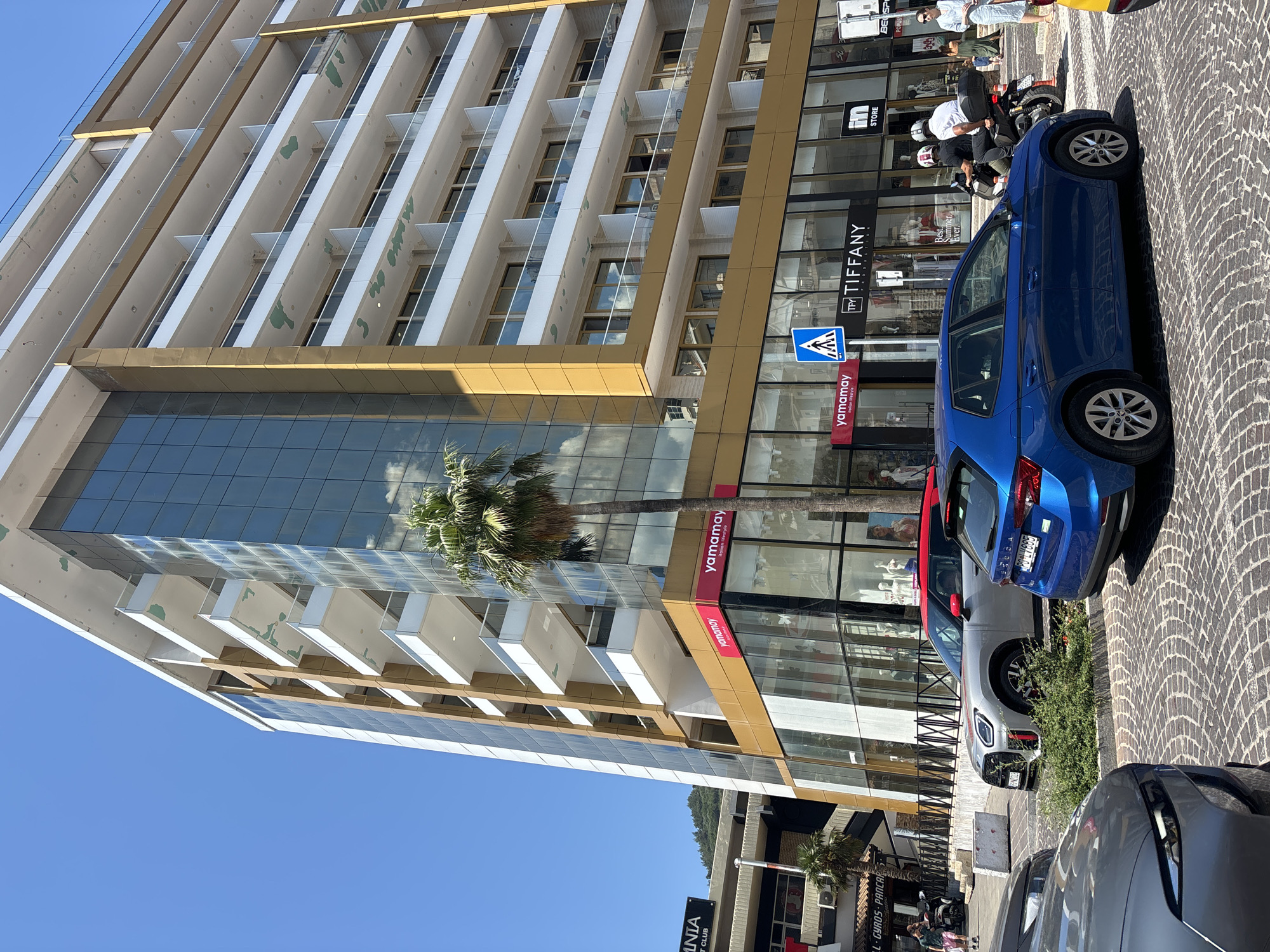 The image shows a street scene in Budva, Montenegro, on a bright, sunny day. A modern, multi-story building with a gold and glass facade dominates the background. The building features multiple balconies with white railings and some appear to have laundry hanging on them. A palm tree stands in front of the building.
In the foreground, there are several cars parked on a cobblestone street. A blue hatchback is in the center, and a silver compact SUV with a red roof is to its left. A dark gray car is partially visible on the far left.
Pedestrians are visible walking on the sidewalk in front of the building, which houses several shops, including "yamamay" and "TIFFANY PRODUCTION". A traffic sign with a pedestrian crossing symbol is also visible. On the right side of the image, two people are riding a scooter, and a woman in a light blue dress is walking on the sidewalk. Another car, yellow, is partially visible on the far right. The overall impression is of a busy commercial area in a Mediterranean city.
