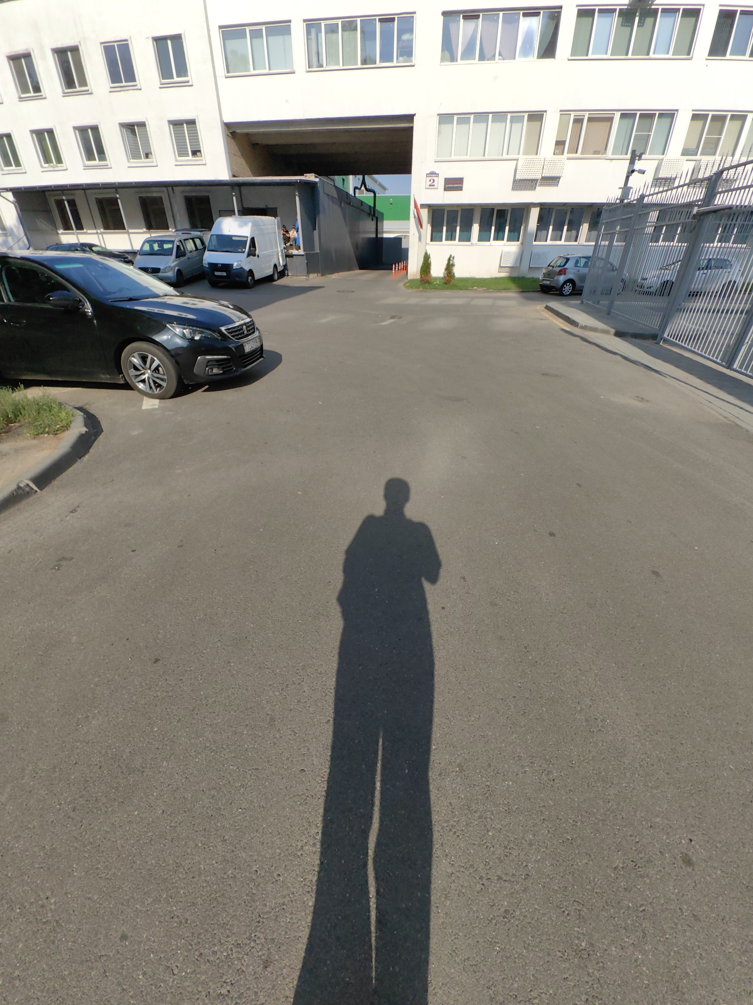 An outdoor, eye-level, wide-angle shot captures a shadow of a person in the foreground on a paved surface, extending from the bottom of the frame upwards. Behind the shadow, to the left, a dark-colored car is parked, facing right. Further back, to the left of the frame, a white van is parked. In the background, a multi-story white building with numerous windows and dark window frames is visible. A metal fence runs along the right side of the frame, parallel to the building. The scene appears to be an industrial or commercial area, possibly a parking lot or access road, with the building suggesting an office or factory complex. The presence of parked vehicles and the architectural style of the building are consistent with urban environments in Minsk, Belarus.