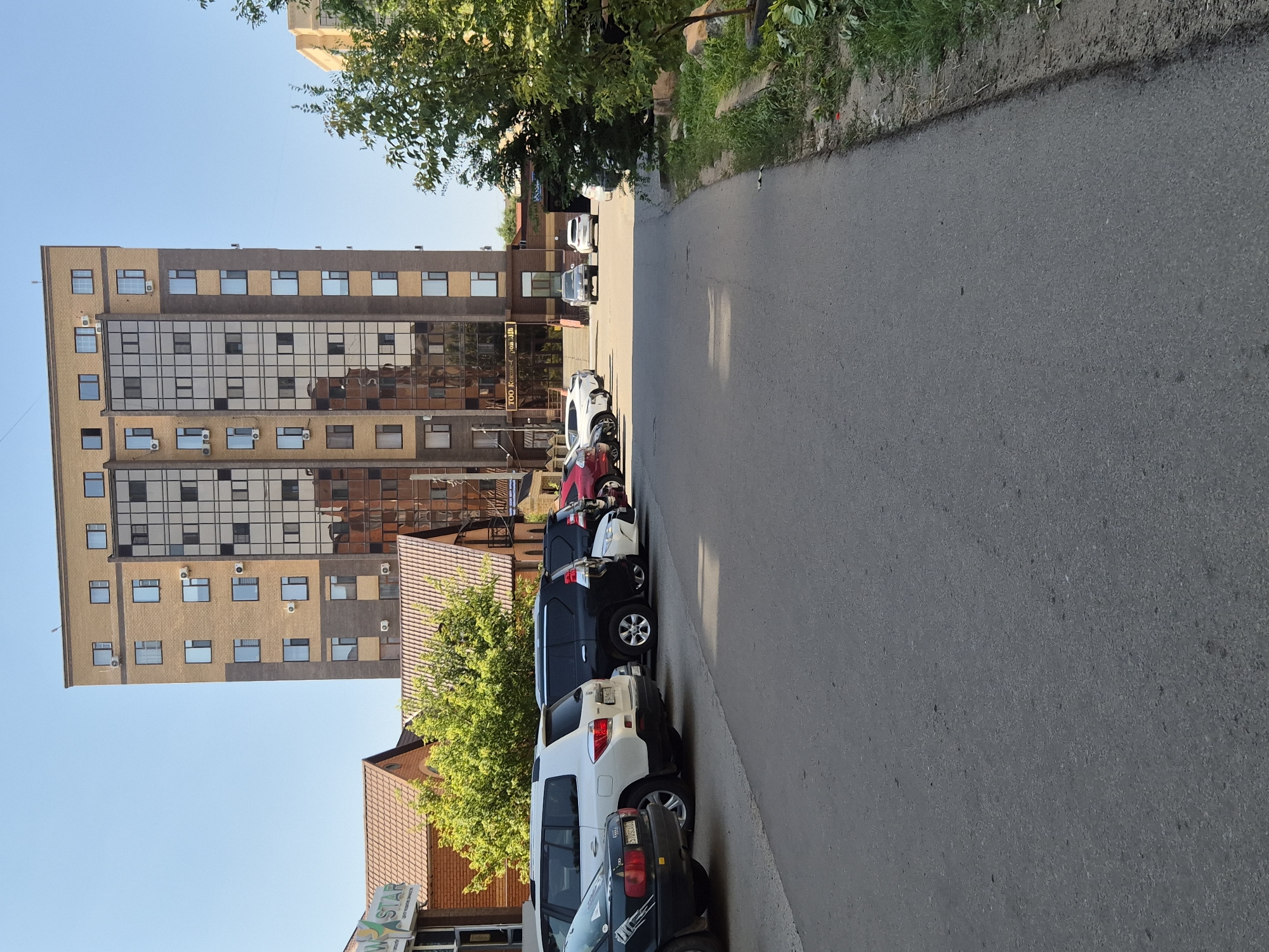 The image shows a street scene in Kokshetau, Kazakhstan, with a tall, modern apartment building dominating the background. The building is a mix of beige brick and dark, reflective glass panels. Several cars are parked on the side of the road in front of the building, including a black Audi, a white SUV, and a black SUV. To the left of the apartment building, there is a lower, reddish-brown building with a gabled roof, partially obscured by trees. The street is paved with asphalt, and there is a strip of grass and foliage along the right side of the road. The sky is clear and blue, indicating a sunny day. There are no people visible in the image. The signage on the apartment building reads "TOO Кокше-Транс-ЛЛБ".