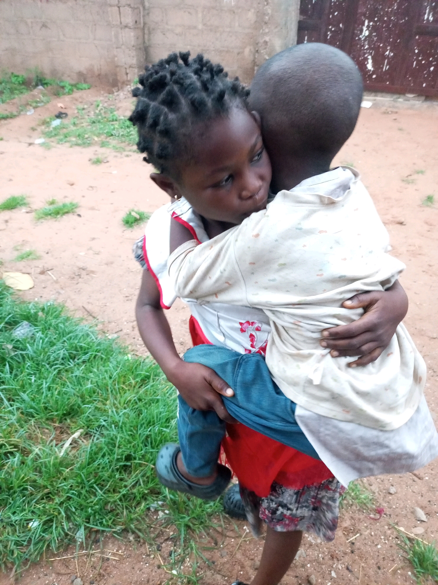Two young children, one seemingly holding the other, are embracing outdoors in a dusty, open area with sparse green vegetation and a brick wall in the background. The child in front, wearing a red skirt and blue pants, is being held by a child with a shaved head. The child being held has their eyes closed and appears to be leaning into the hug. The scene is set in Tempure, Nigeria.
