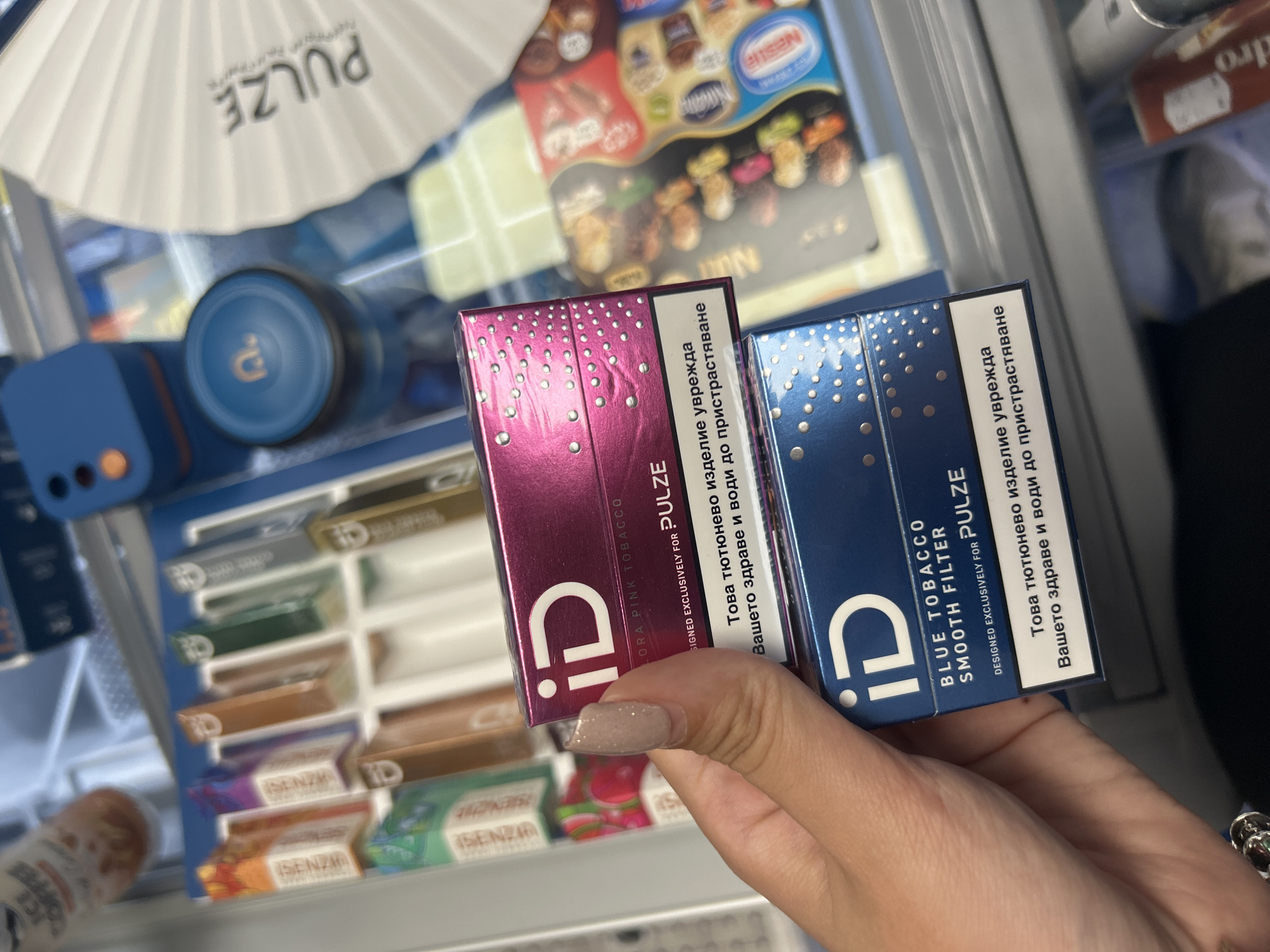 This is a close-up, eye-level shot of a person's hands holding two packs of what appear to be tobacco products. The packs are magenta and blue, decorated with white dots. The magenta pack has the text "ID" and "LORA PINK TOBACCO" printed on it, along with a warning in Cyrillic script. The blue pack has "ID" and "BLUE TOBACCO SMOOTH FILTER" printed on it, also with a Cyrillic warning.
In the background, a white, fan-like object with the text "PULZE" is visible. Below this, a blue cylindrical object with a circular indicator and an orange button is present. Further in the background, a display case filled with various colored packets, likely containing similar tobacco products, is visible. Some of these packets are labeled "SENZIA". A partial view of a beverage container, possibly an iced coffee, is also visible in the lower left corner. The overall setting appears to be a retail environment, possibly a convenience store or a vending machine area. The lighting suggests it is indoors. The focus is on the tobacco products in the foreground, held by the person's hands, one of which has neatly manicured fingernails.