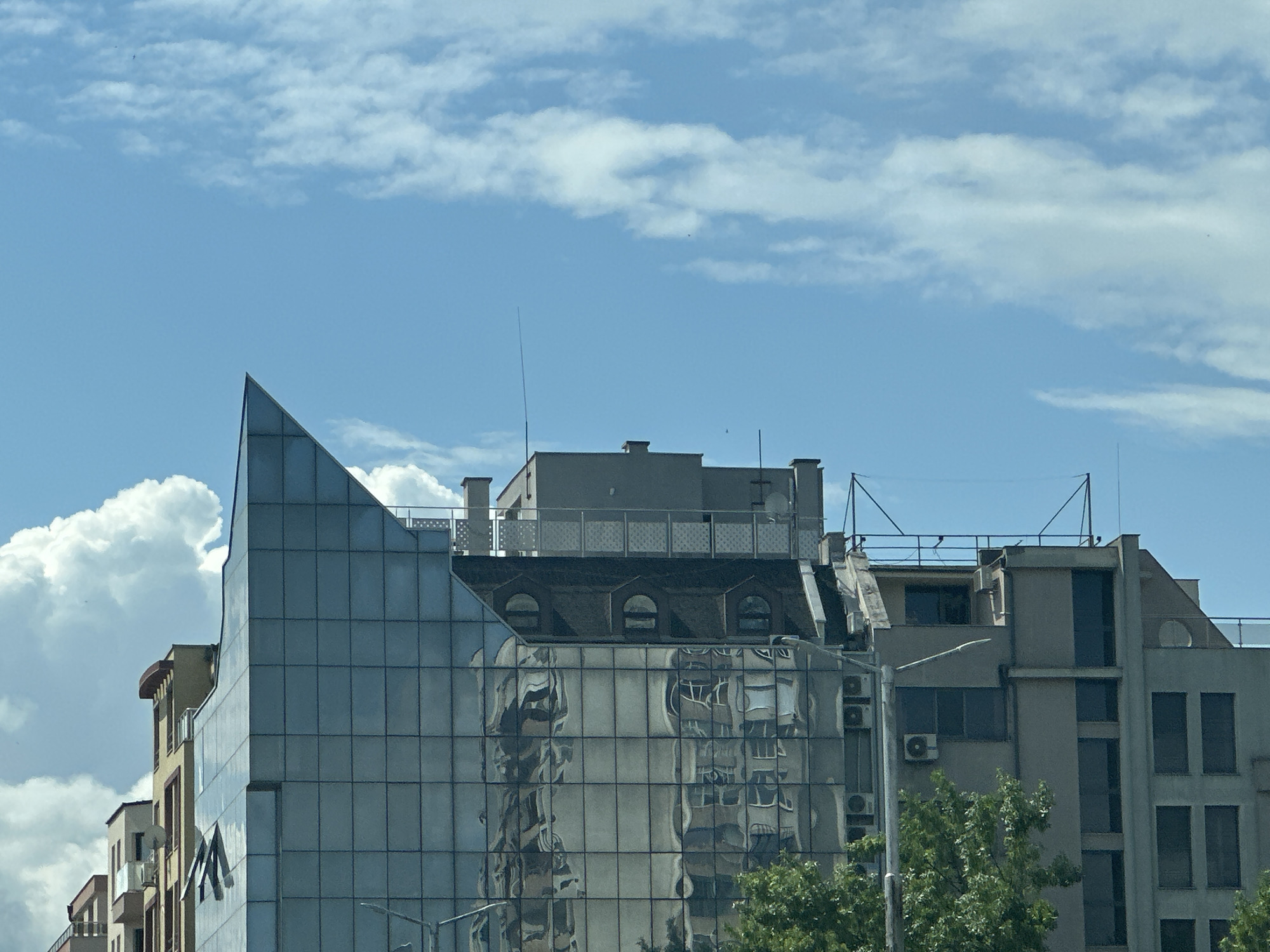 The image shows a partial view of several buildings in Plovdiv, Bulgaria, under a blue sky with scattered white clouds. The most prominent building features a glass facade with a distinct triangular peak, reflecting the image of another building. Adjacent to the glass building is a building with a rooftop terrace. To the right is a concrete building with visible windows, air conditioning units, and antennas. Trees are in the foreground obscuring a portion of the buildings. The buildings appear to be located in an urban area.