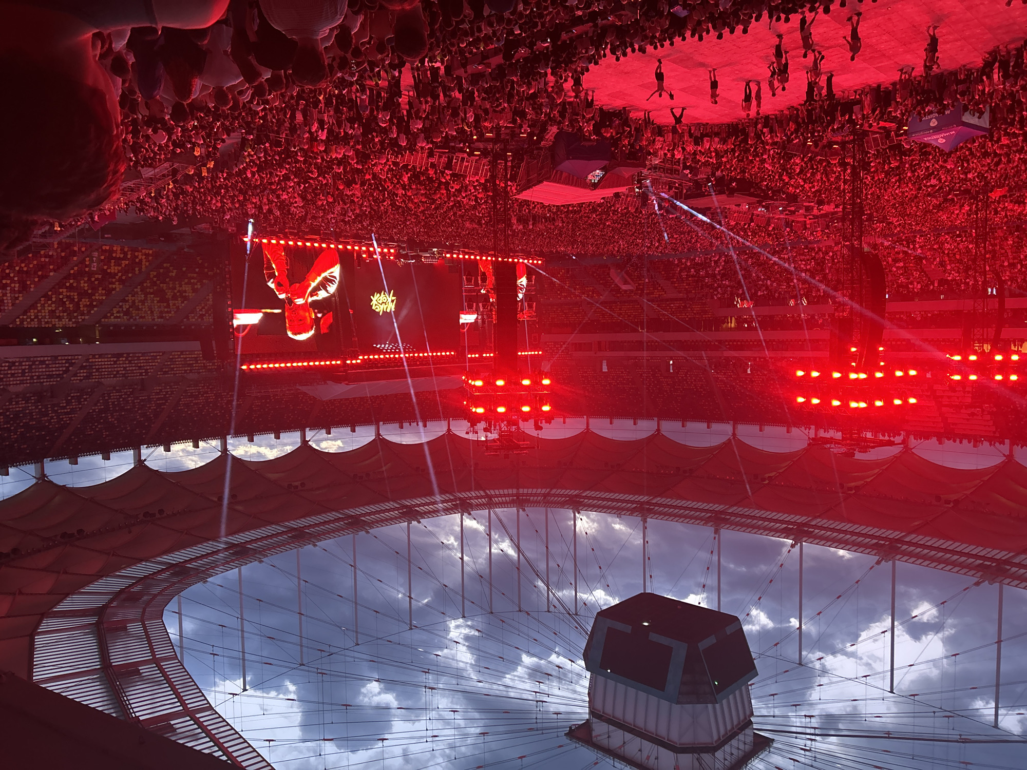 The image shows an indoor stadium at a concert with a large crowd of people. The venue has a partially covered roof, with a structure at the center. The concert stage features a large screen displaying images and stage lighting, which is predominantly red. Figures can be seen on the stage. The crowd fills the seating areas and floor of the stadium.