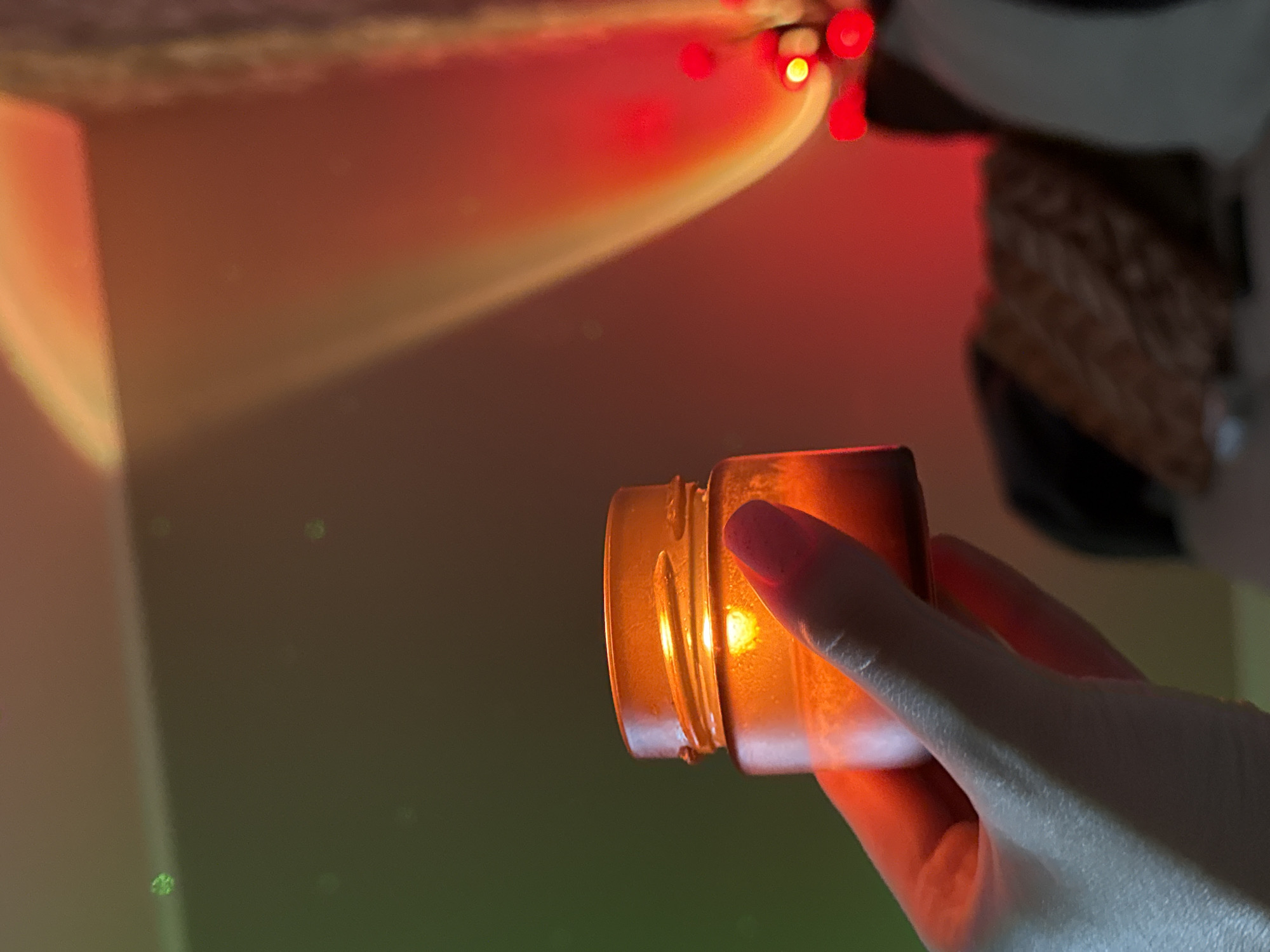 A hand with pink nail polish holds an amber glass jar in front of a wall illuminated by red and green lights. The jar is small and appears to be a candle or a container for a cosmetic product. The light creates a soft glow around the jar. In the background, there are blurred objects and abstract light patterns. The overall impression is one of a cozy, ambient setting.