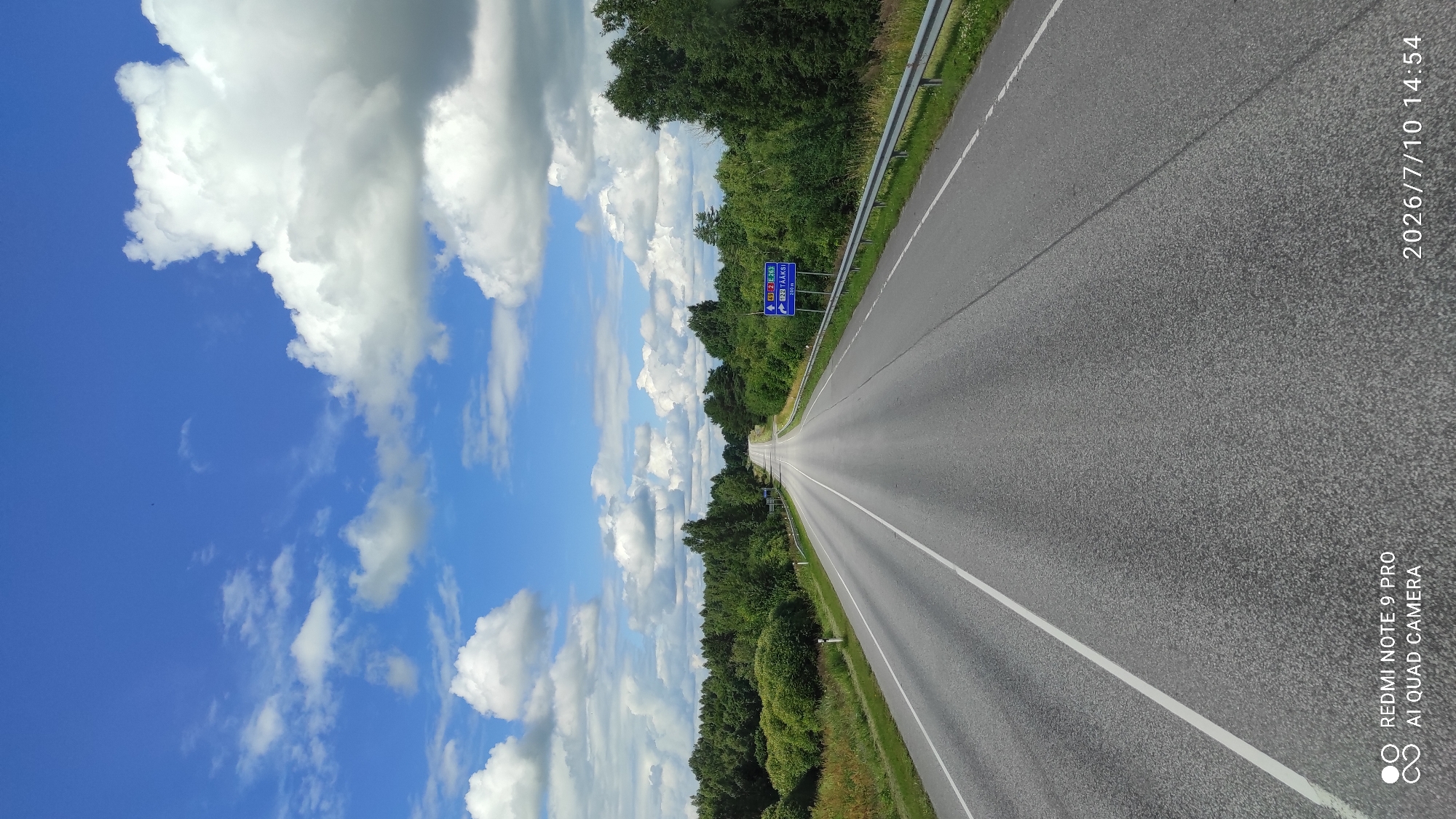 The image shows a straight, paved road with double white lines in the center, leading into the distance. The road is flanked by lush green trees and vegetation on both sides, with a metal guardrail on the right. The sky is a vibrant blue with scattered white cumulus clouds. In the distance, on the right side of the road, a blue traffic sign is visible, indicating directions and distances to various locations. The sign appears to show route numbers E263 and E77, with text likely in Estonian. The timestamp on the image reads "2026/7/10 14:54", and the text "REDMI NOTE 9 PRO AI QUAD CAMERA" is present in the lower left corner, indicating the device used to capture the photo and its placement within the city of Võivaku, Estonia.